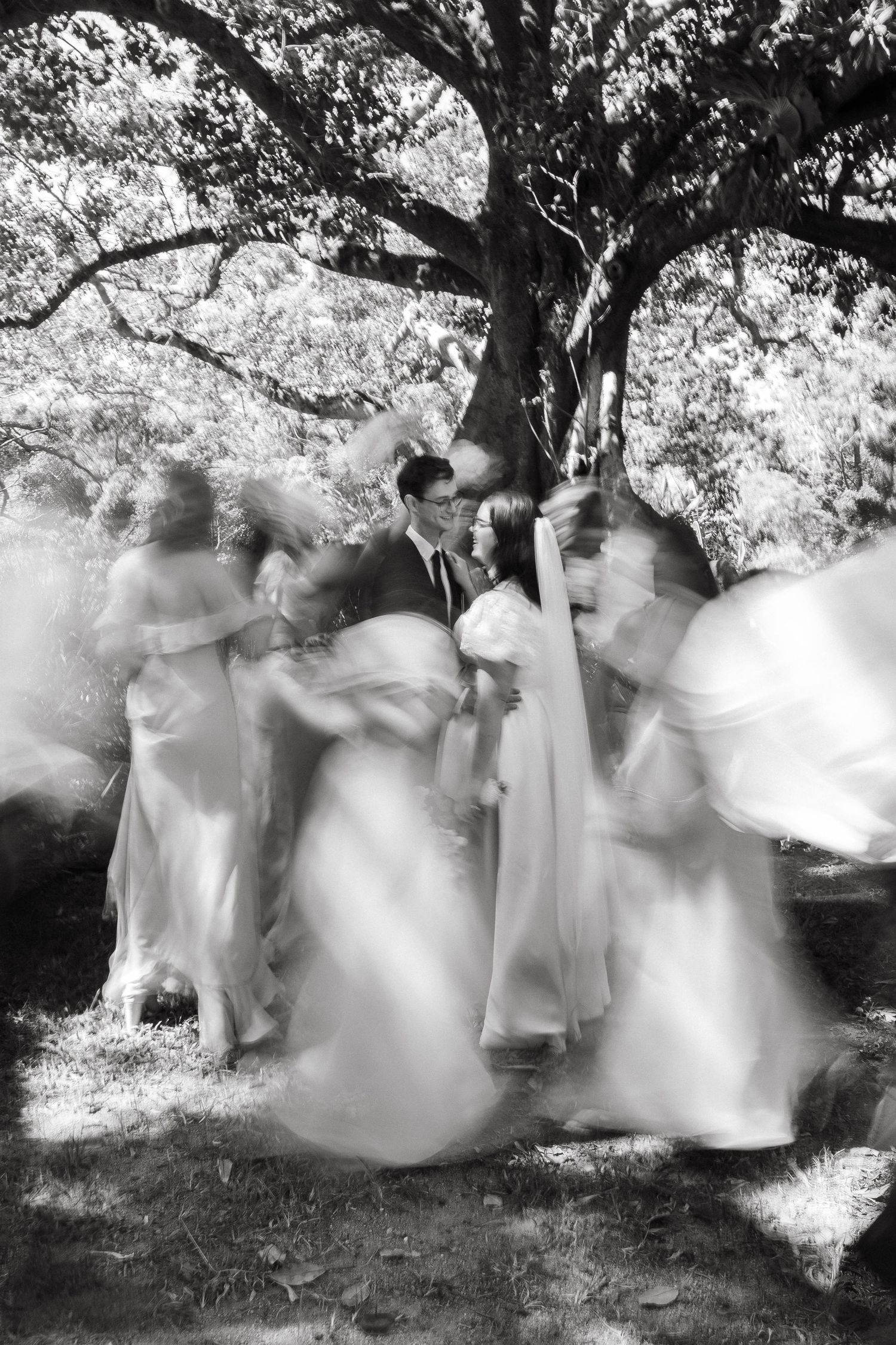 Black and white photo of a wedding party dancing under a large tree, with motion blur creating a dreamy effect.