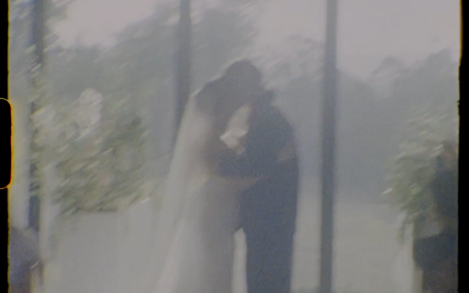 A couple shares a romantic first kiss at their wedding ceremony, framed by lush greenery in a dreamy, film-style photo.