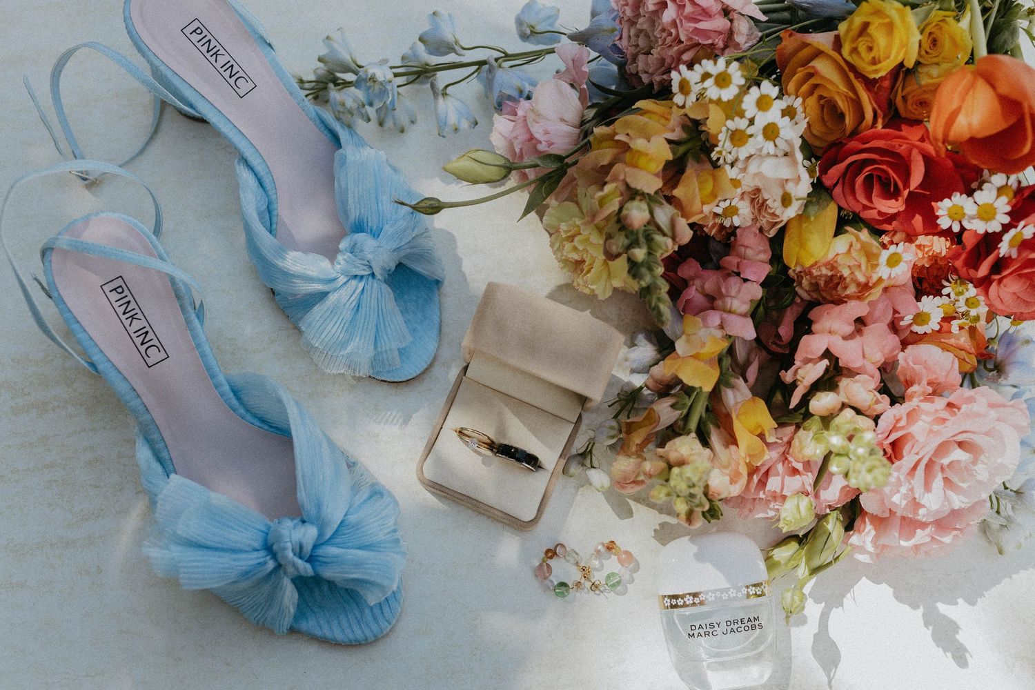 Blue bow heels, wedding rings in box, and colorful bridal bouquet with roses and daisies on white surface.