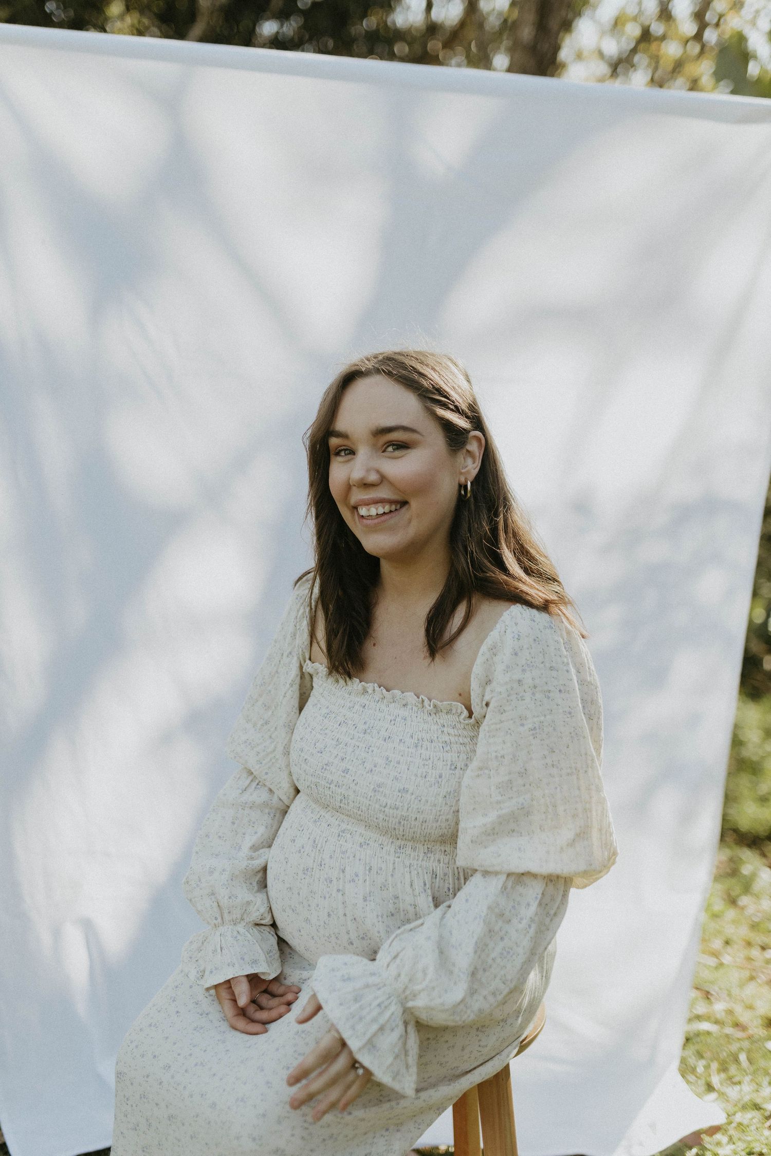 Happy pregnant woman in cream floral smocked dress smiling outdoors against white fabric backdrop.