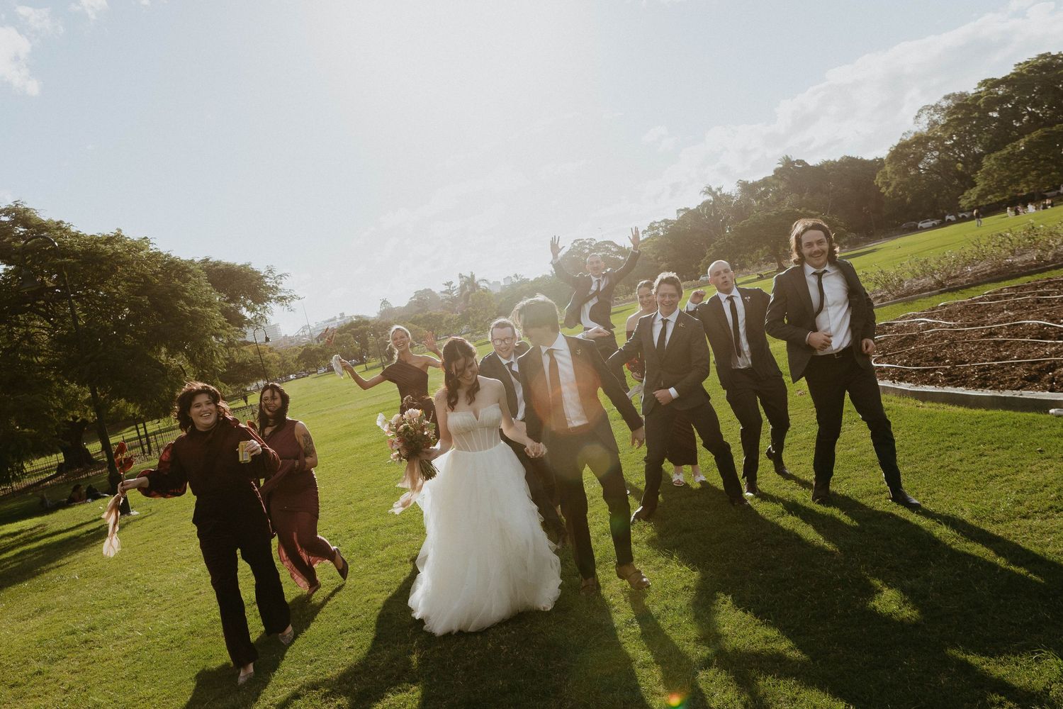Wedding party celebrating outdoors on lush green lawn with bride in white gown and groom in sunny setting.