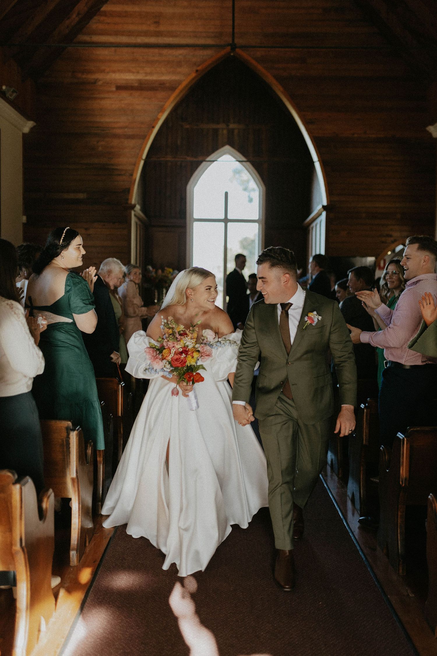 Newlyweds walking down aisle of rustic wooden chapel holding colorful bridal bouquet.