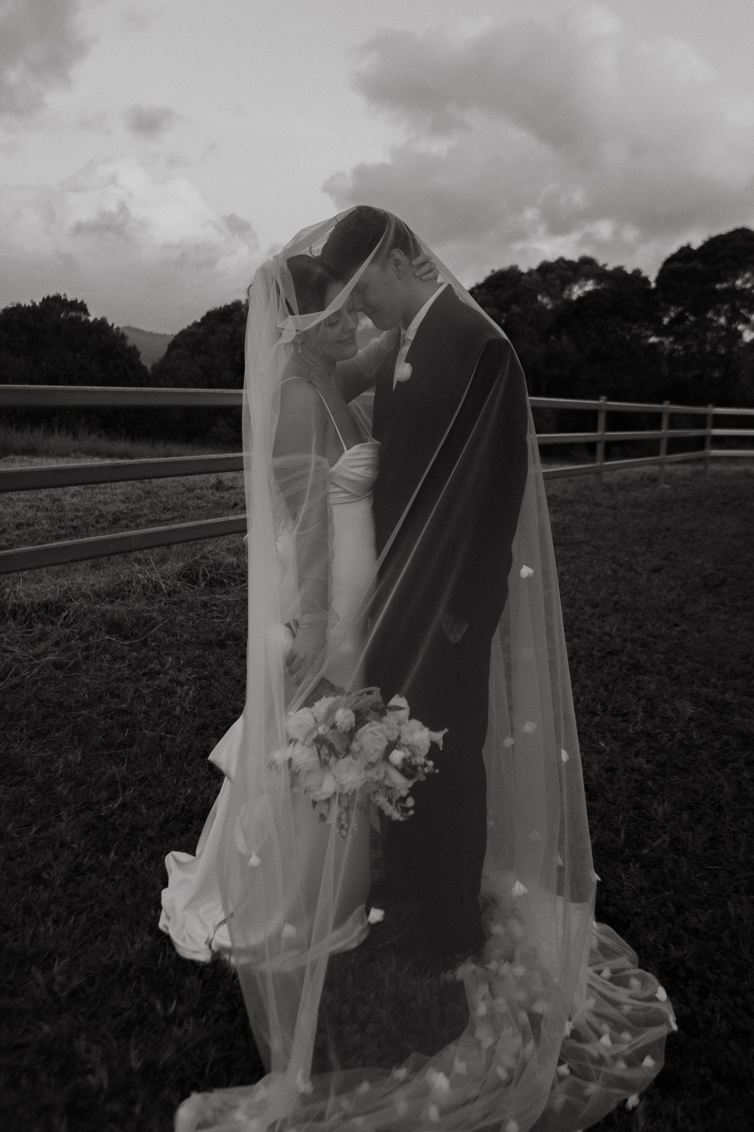 Black and white photo of bride and groom kissing under wedding veil outdoors, holding bouquet near wooden fence.