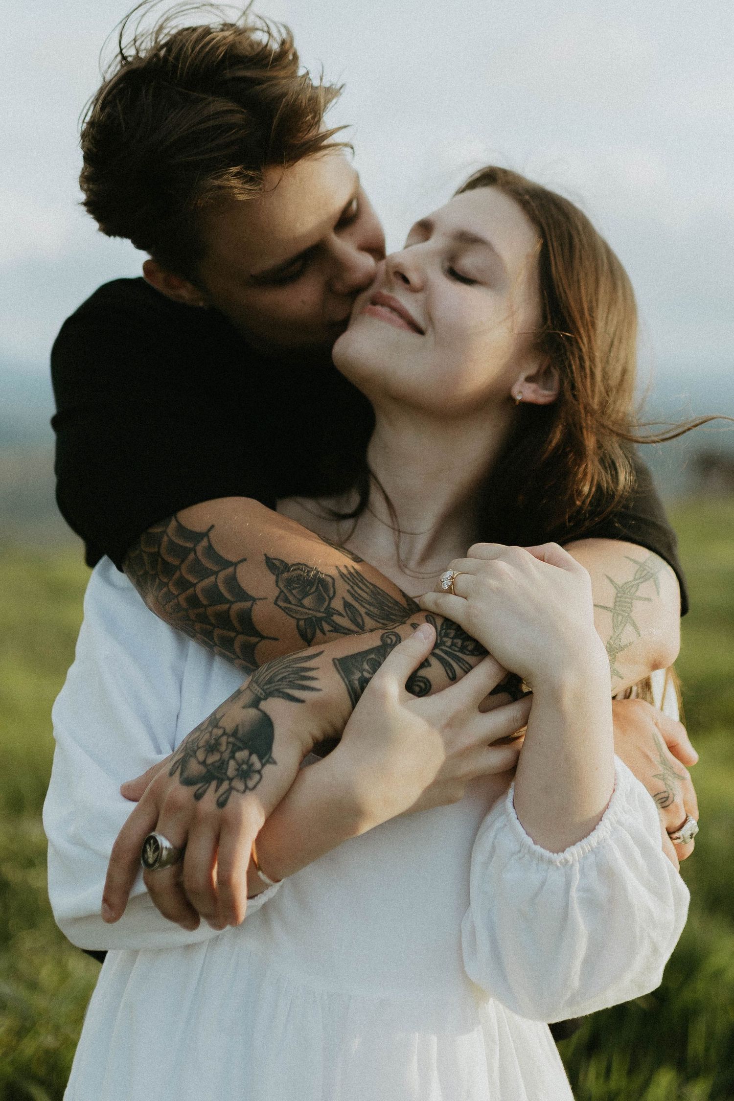 A man with tattooed arms embraces a woman in a white dress from behind, kissing her cheek outdoors.