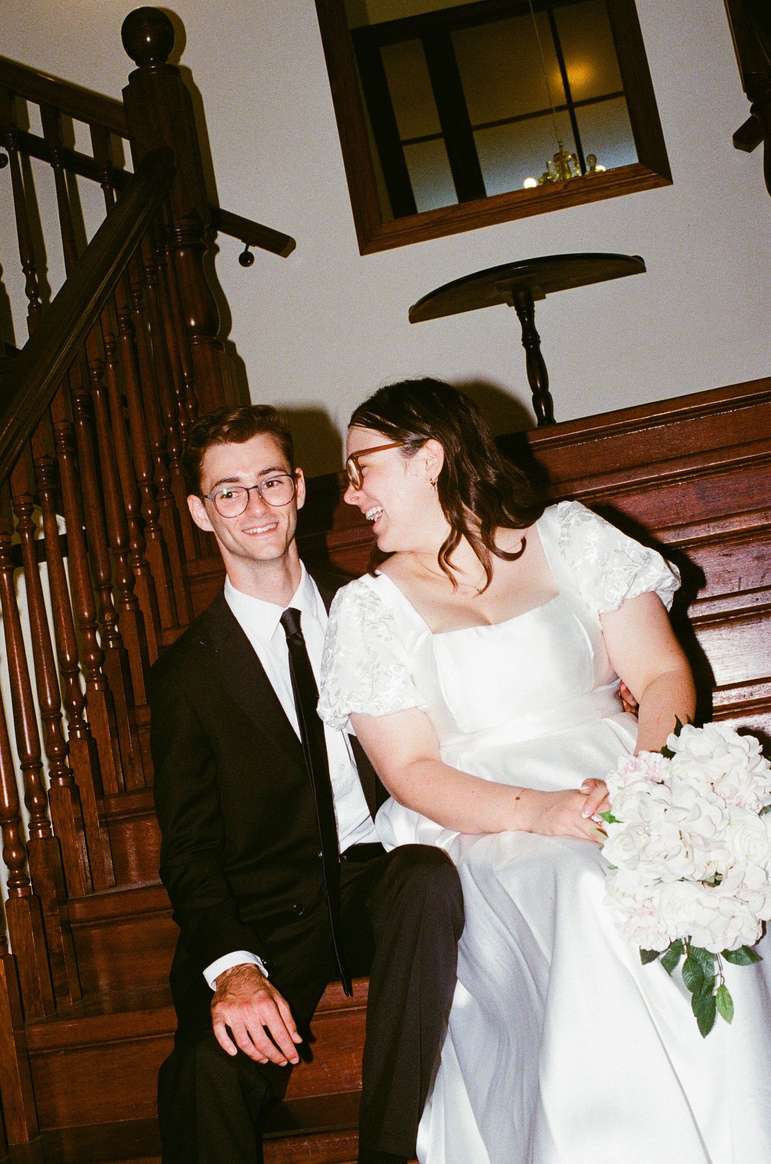Bride and groom sitting on wooden staircase smiling at each other, bride holding white floral bouquet.