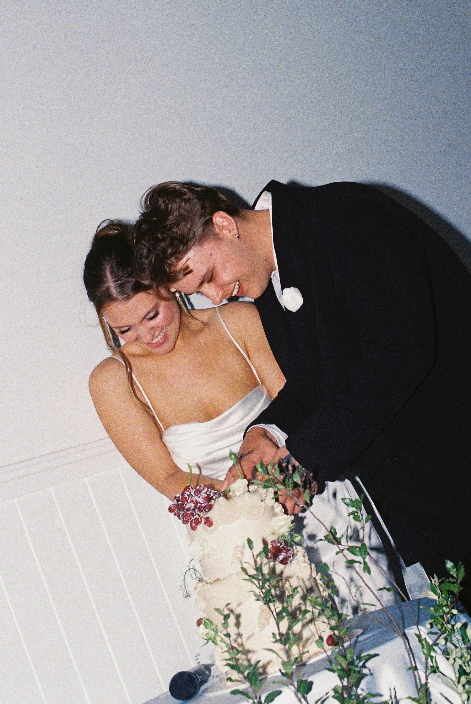 A smiling bride and groom in formal attire cutting their white wedding cake adorned with red berries and greenery.