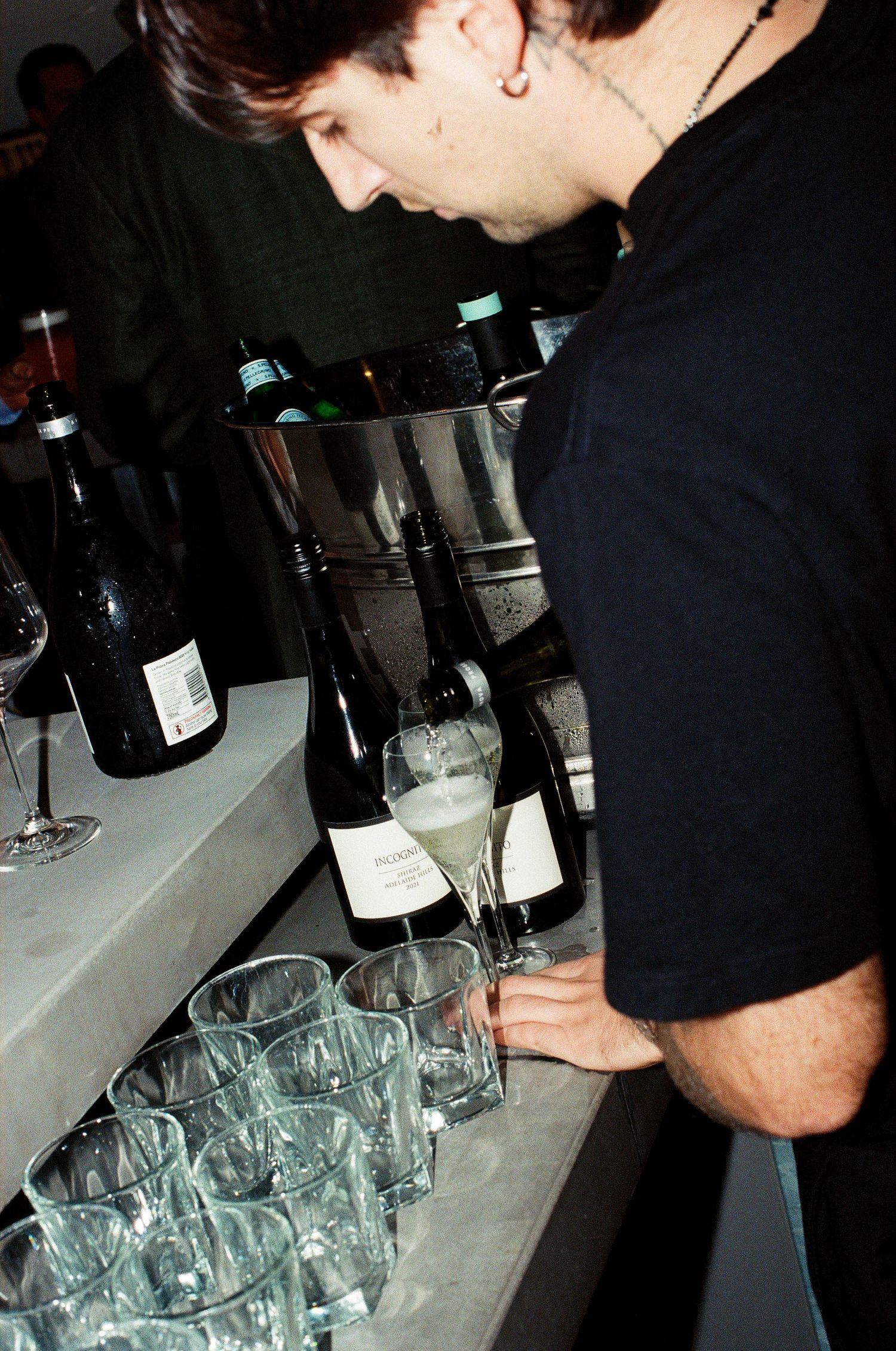 Bartender pouring white wine into glasses at an event, with multiple wine bottles chilling in an ice bucket.