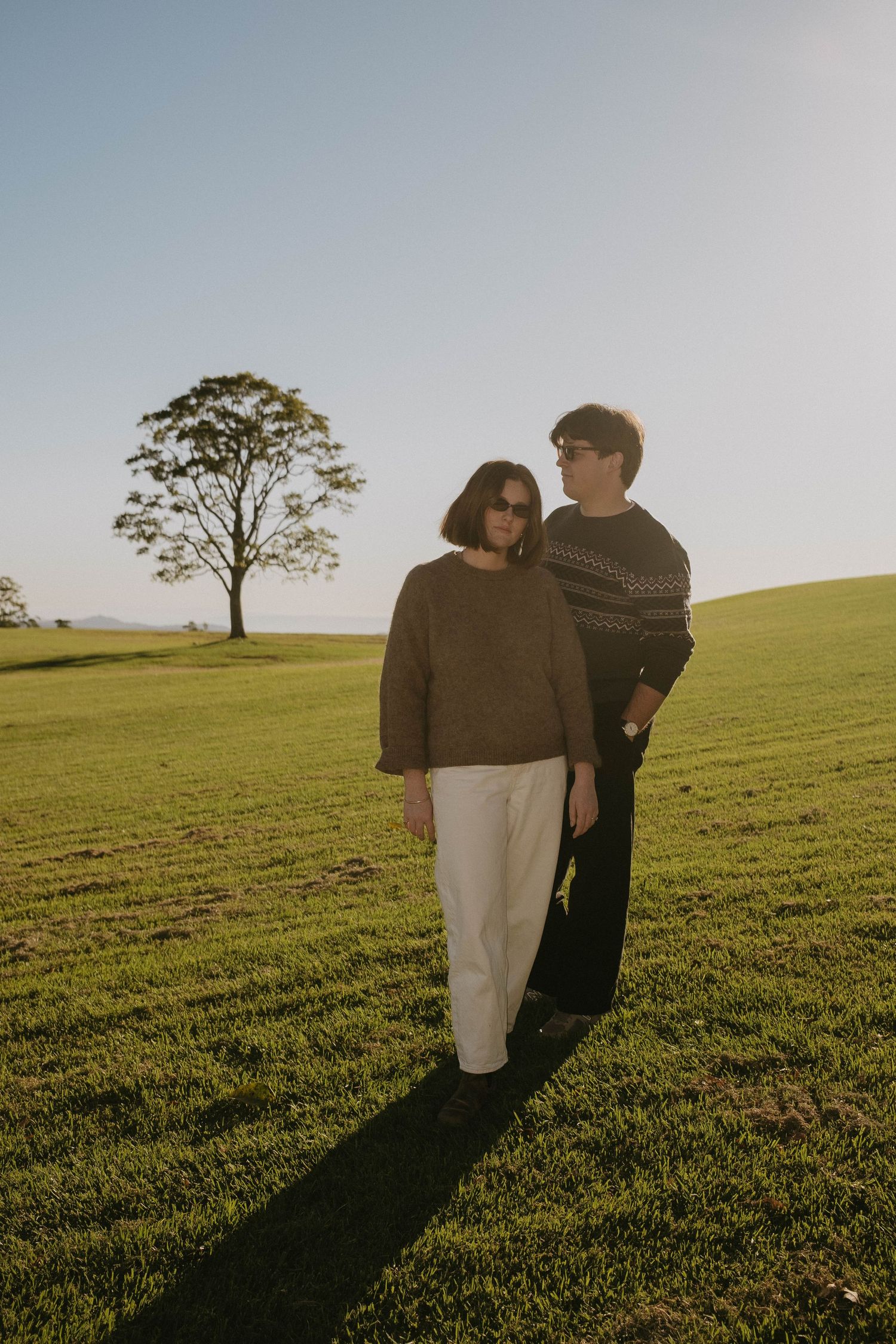 Couple standing in a green field at golden hour with a lone tree in the background under a clear sky.