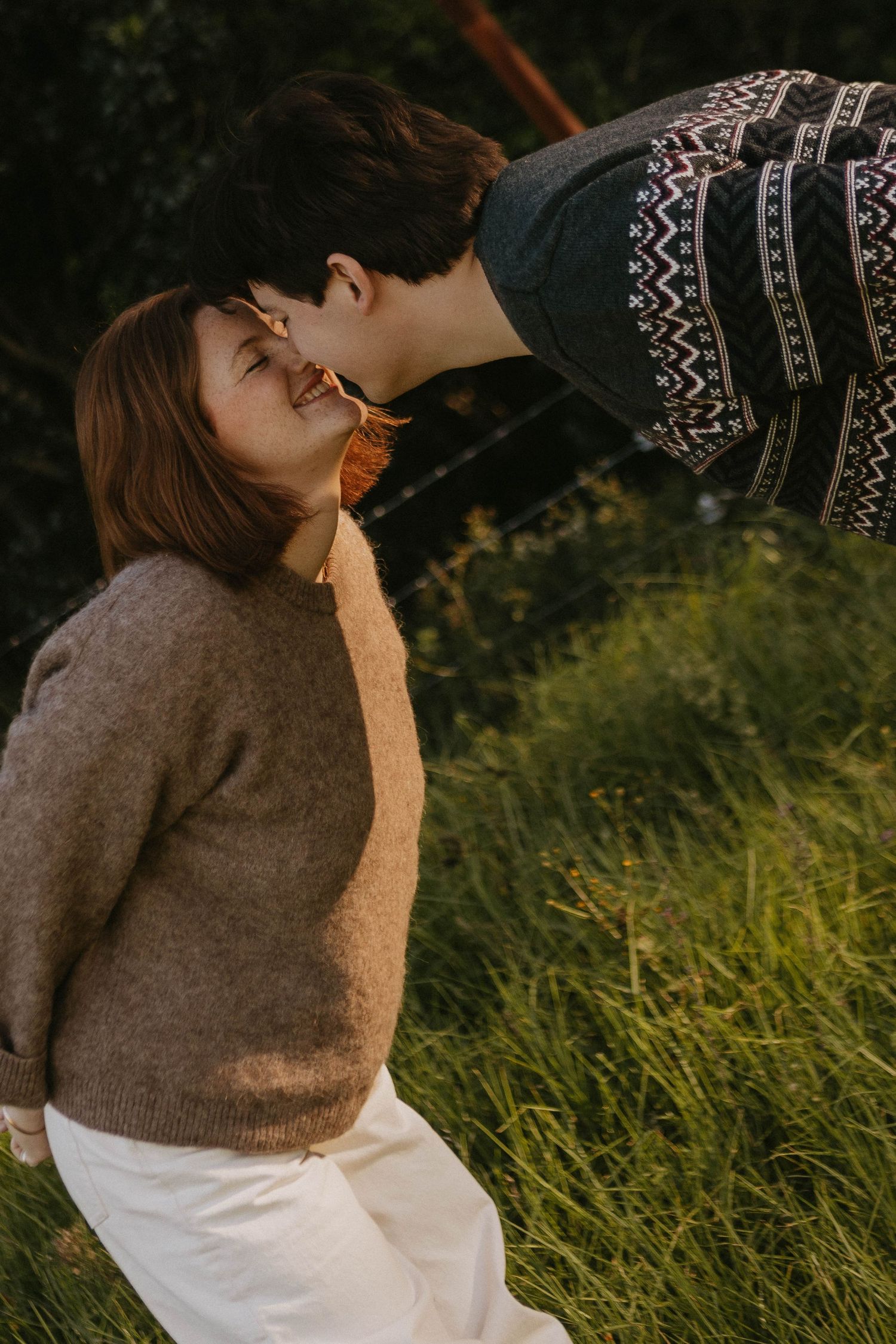 A couple shares a tender moment outdoors, man leaning to kiss woman's forehead near lush green grass.