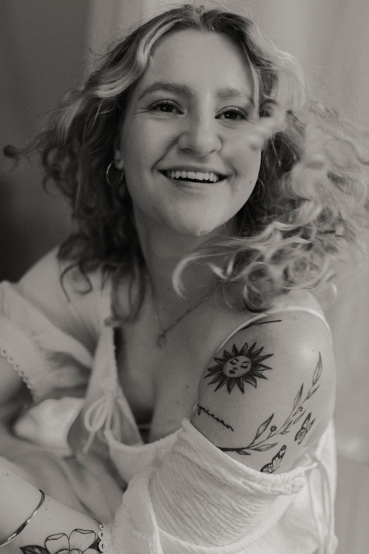Black and white photo of smiling woman with curly hair and sun tattoo on shoulder.