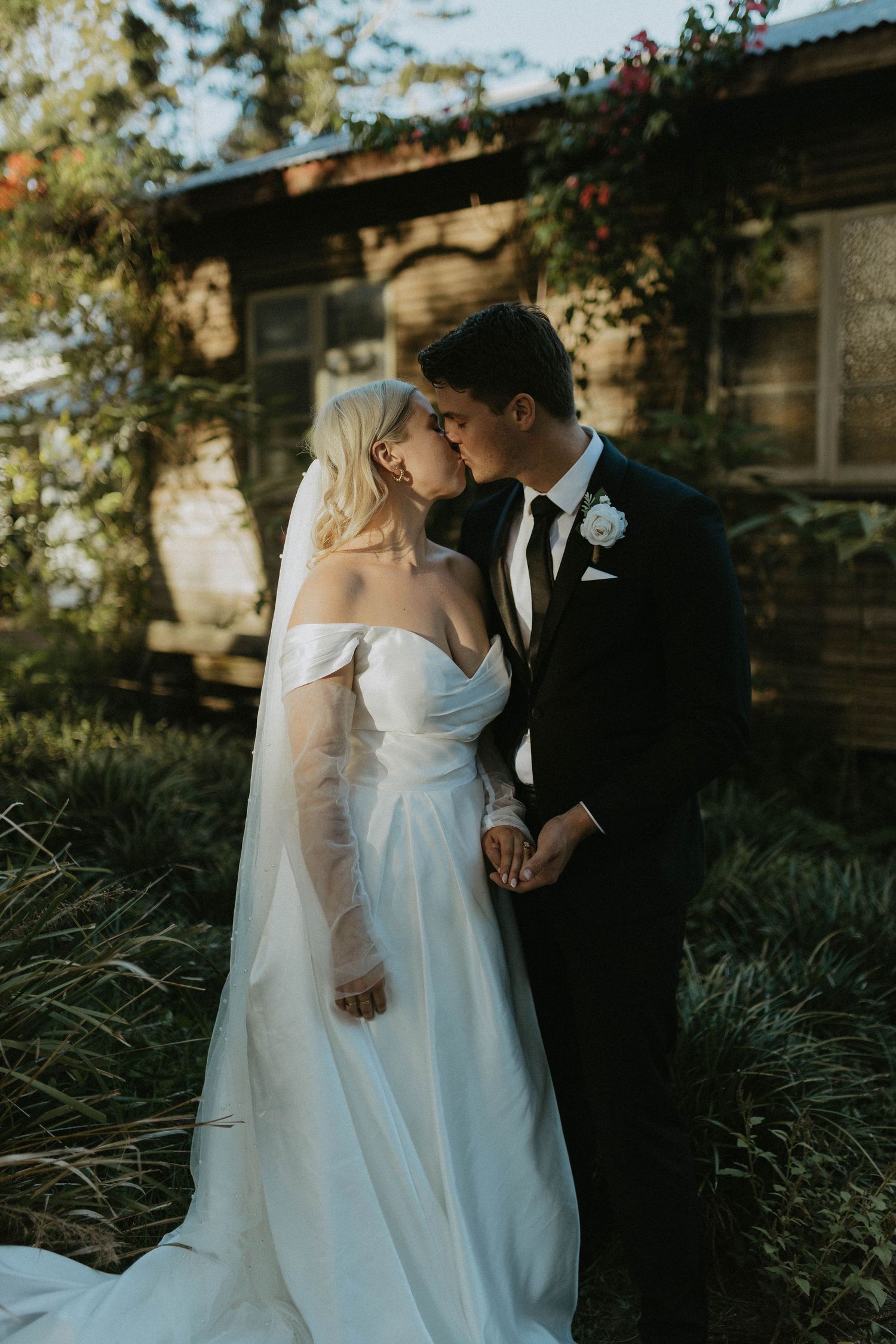 Bride and groom sharing a kiss in garden at golden hour, bride in off-shoulder gown.