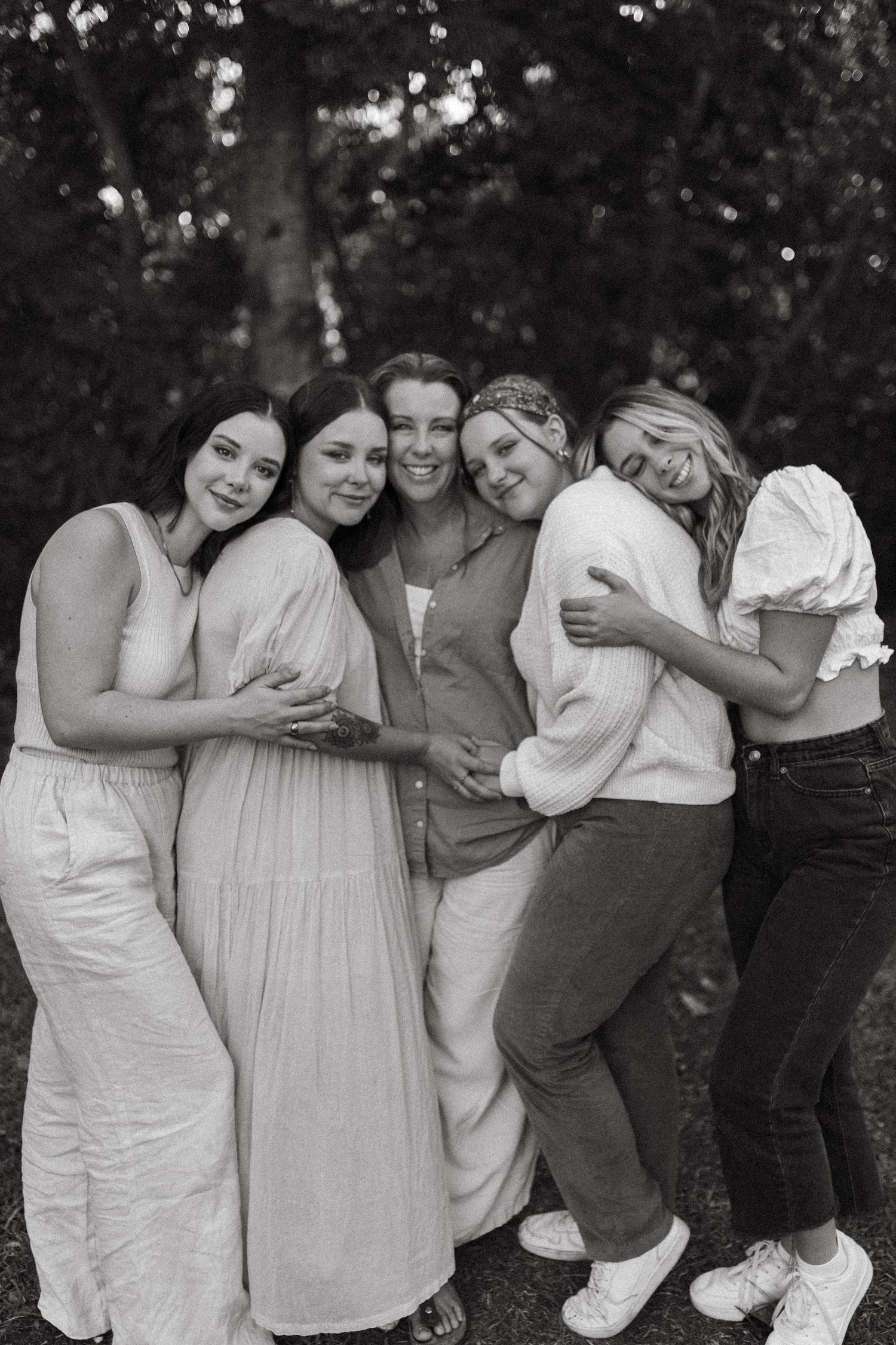 Black and white portrait of five women smiling and embracing each other warmly in an outdoor wooded setting.