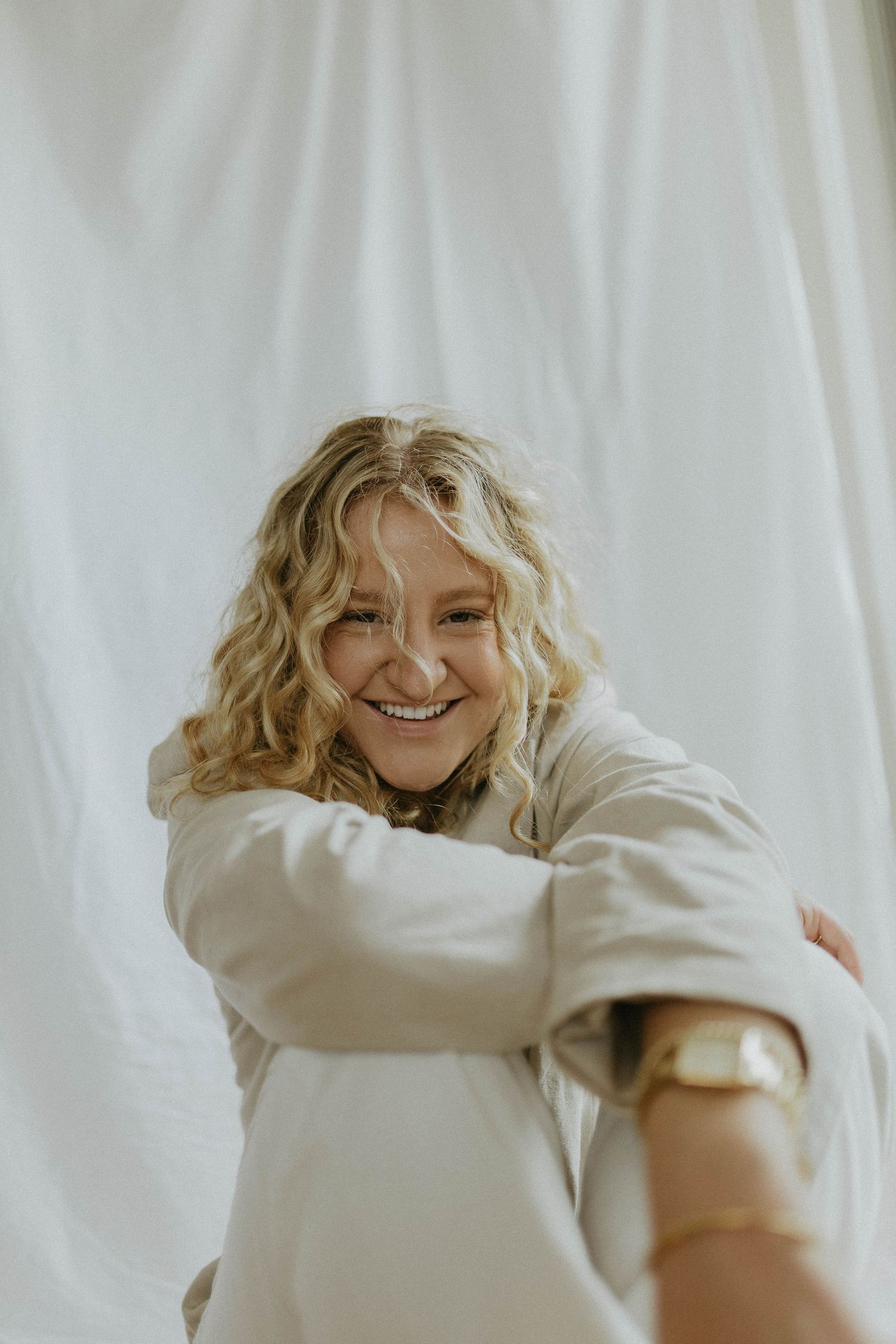Laughing blonde curly-haired woman in beige oversized shirt with gold watch against white curtain.