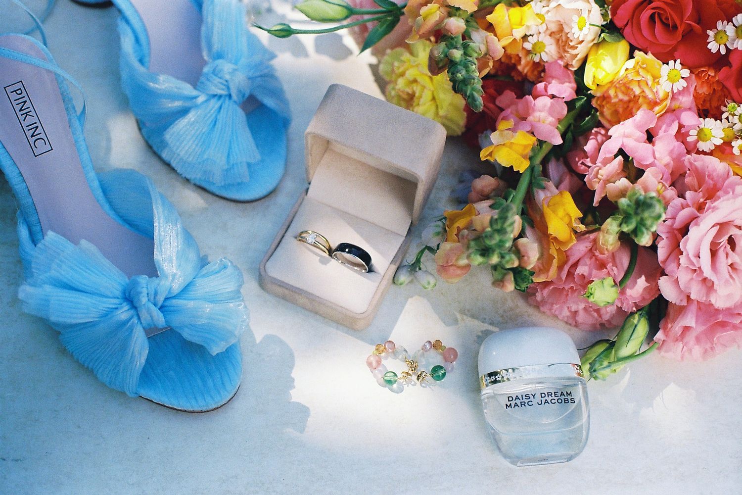 Blue bow wedding heels, ring box with bands, and colorful floral bouquet on blue surface.