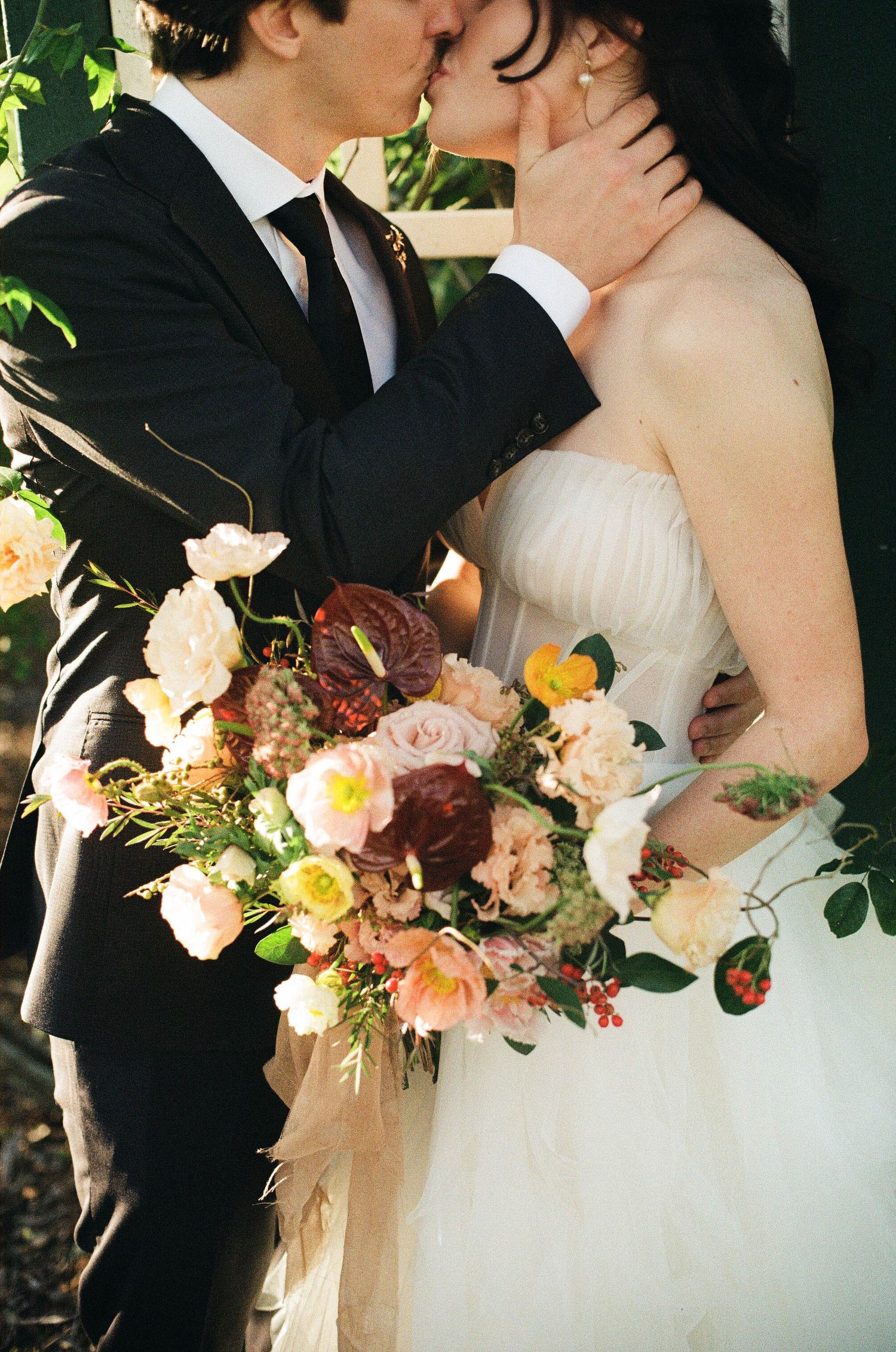 Bride and groom sharing a kiss, bride holding a lush bouquet of pink, burgundy, and yellow flowers.