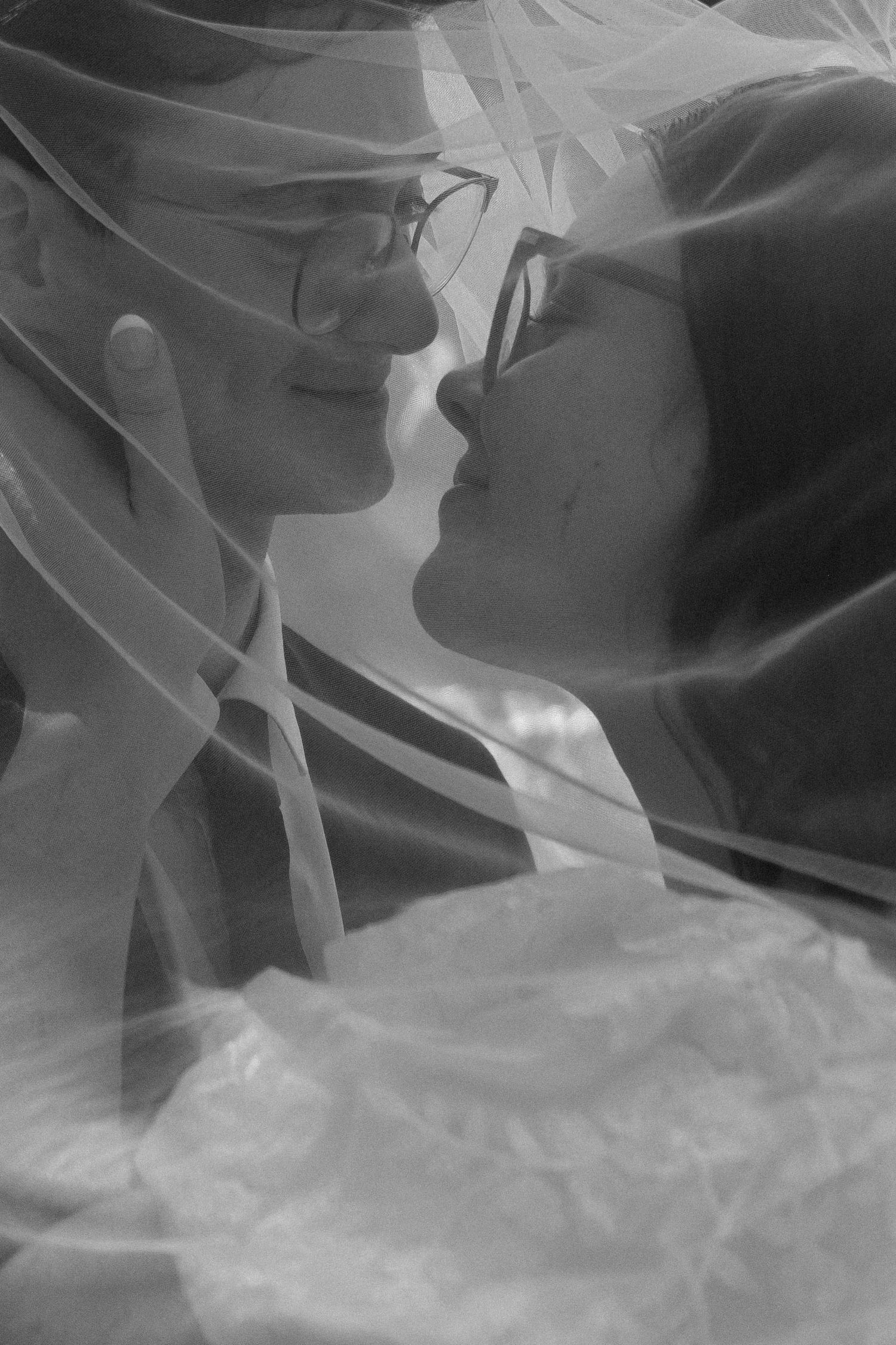 Black and white photo of a couple nearly kissing under a bridal veil, captured in an intimate wedding moment.