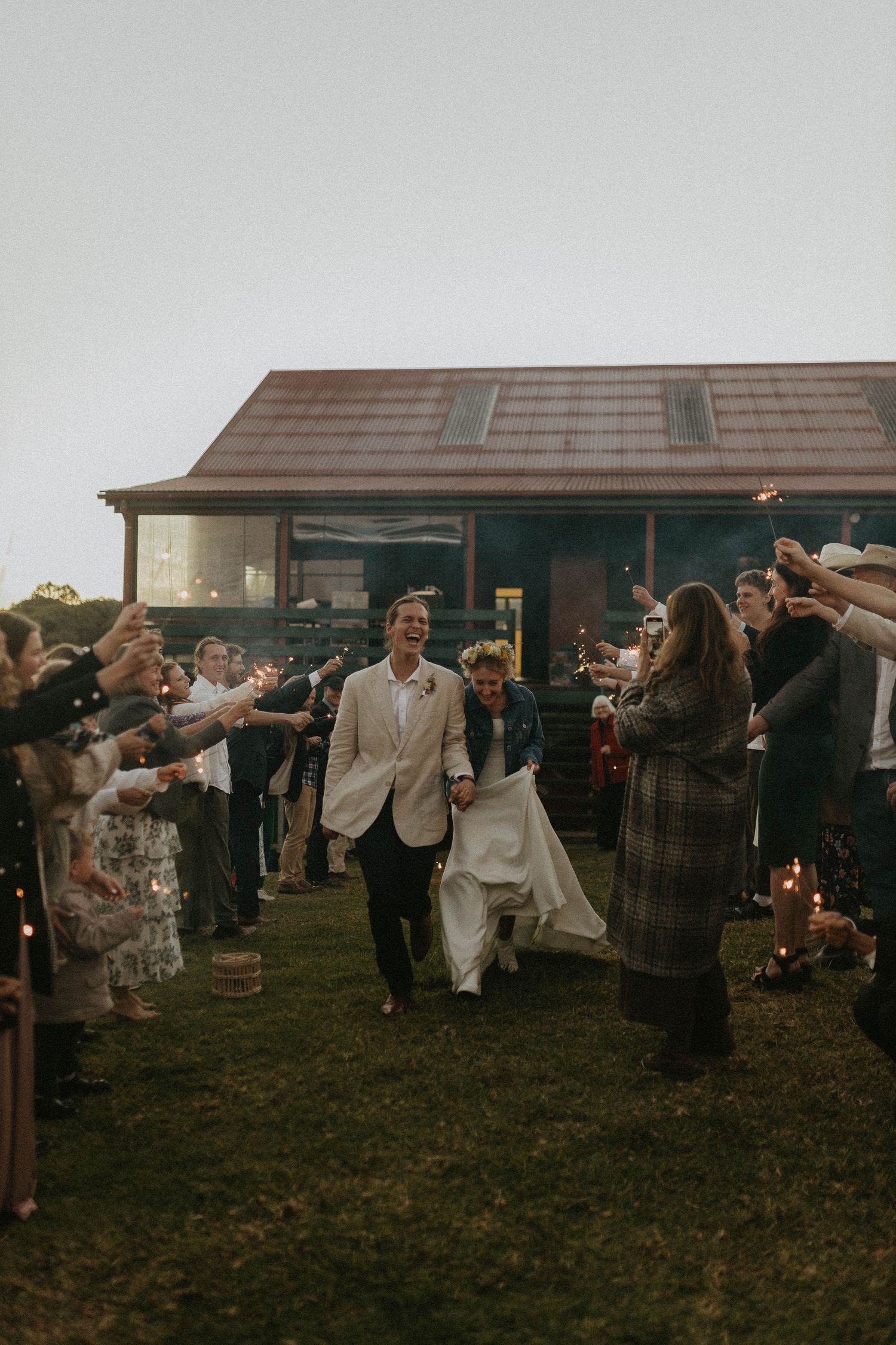 Bride and groom exit wedding reception through sparkler tunnel surrounded by cheering guests at twilight.