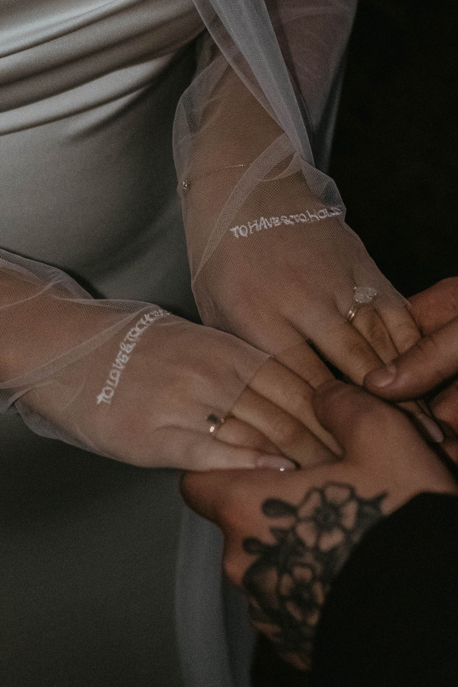 Close-up of two hands holding, one wearing a diamond ring and sheer embroidered sleeve, the other with a floral tattoo.