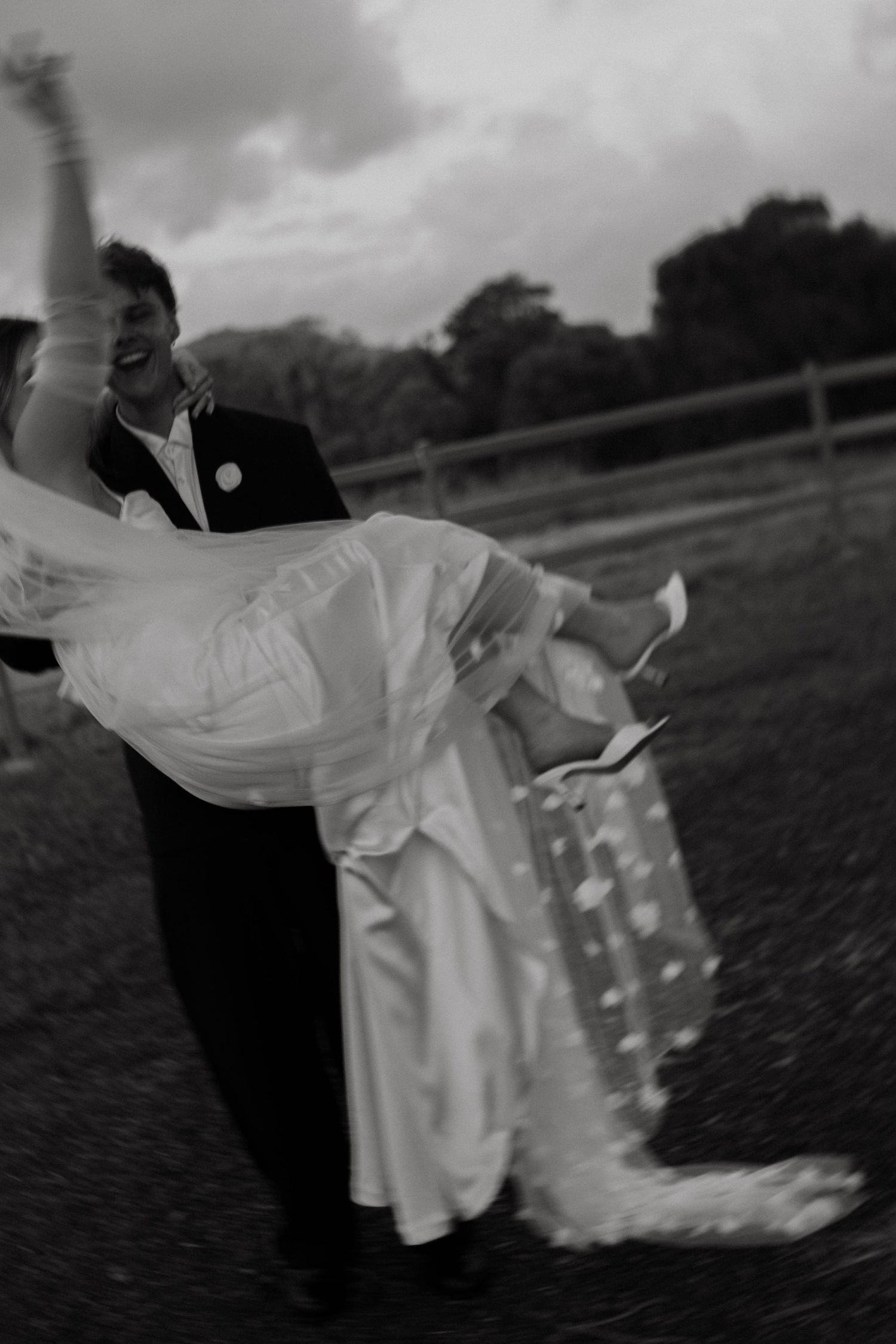 Groom carries bride in his arms outdoors in a joyful black and white wedding photo.