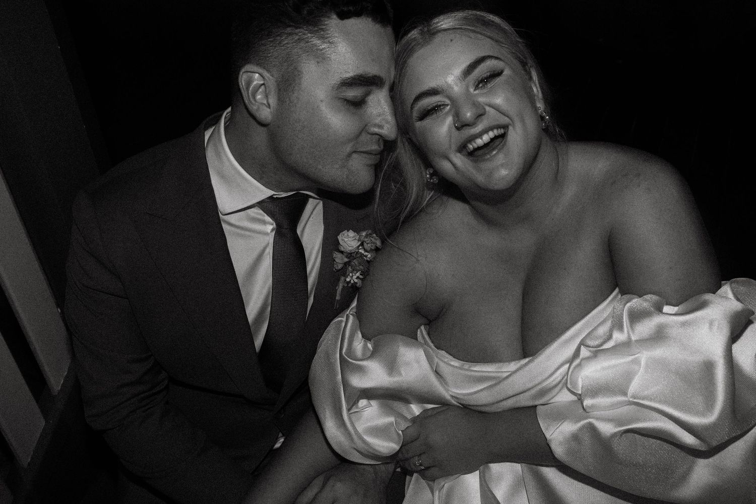Black and white photo of groom kissing laughing bride in satin off-shoulder gown.