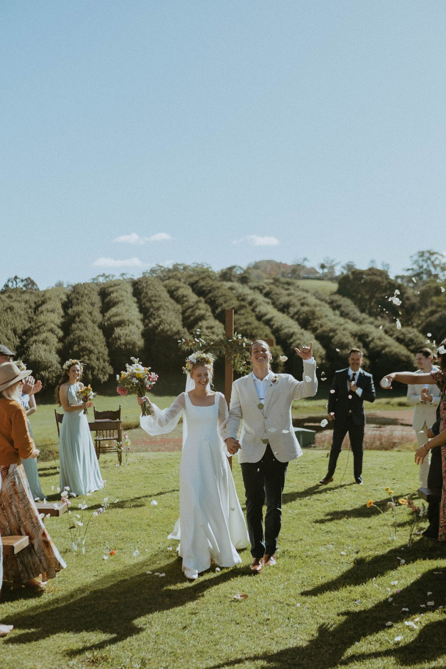 Newlyweds walk down aisle as guests toss confetti at sunny outdoor wedding ceremony on green lawn.