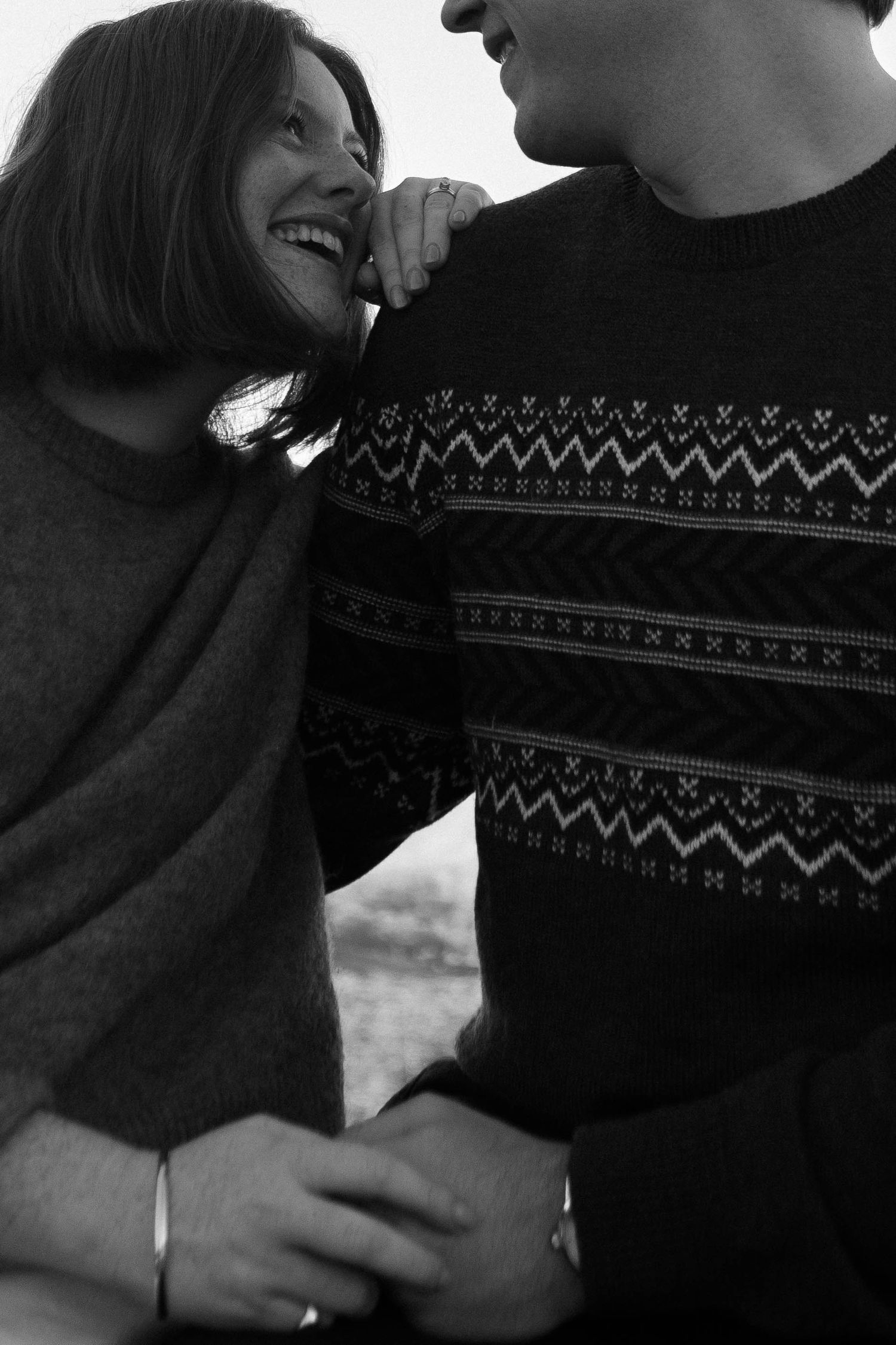 Black and white photo of a laughing woman and man in a Fair Isle sweater sharing a joyful embrace outdoors.