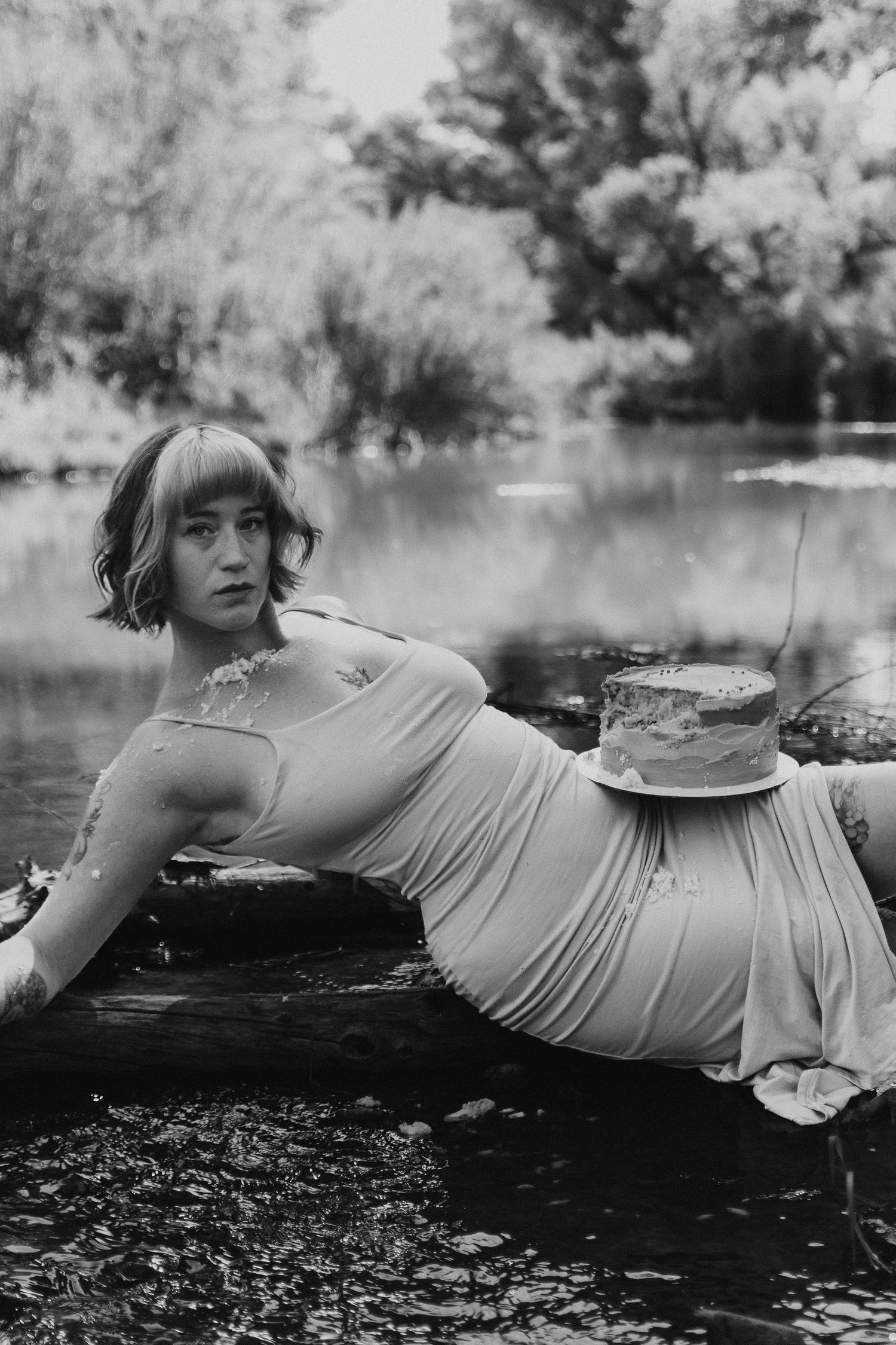 A person in a light-colored dress lies on a dark surface near water in a black and white artistic photograph.