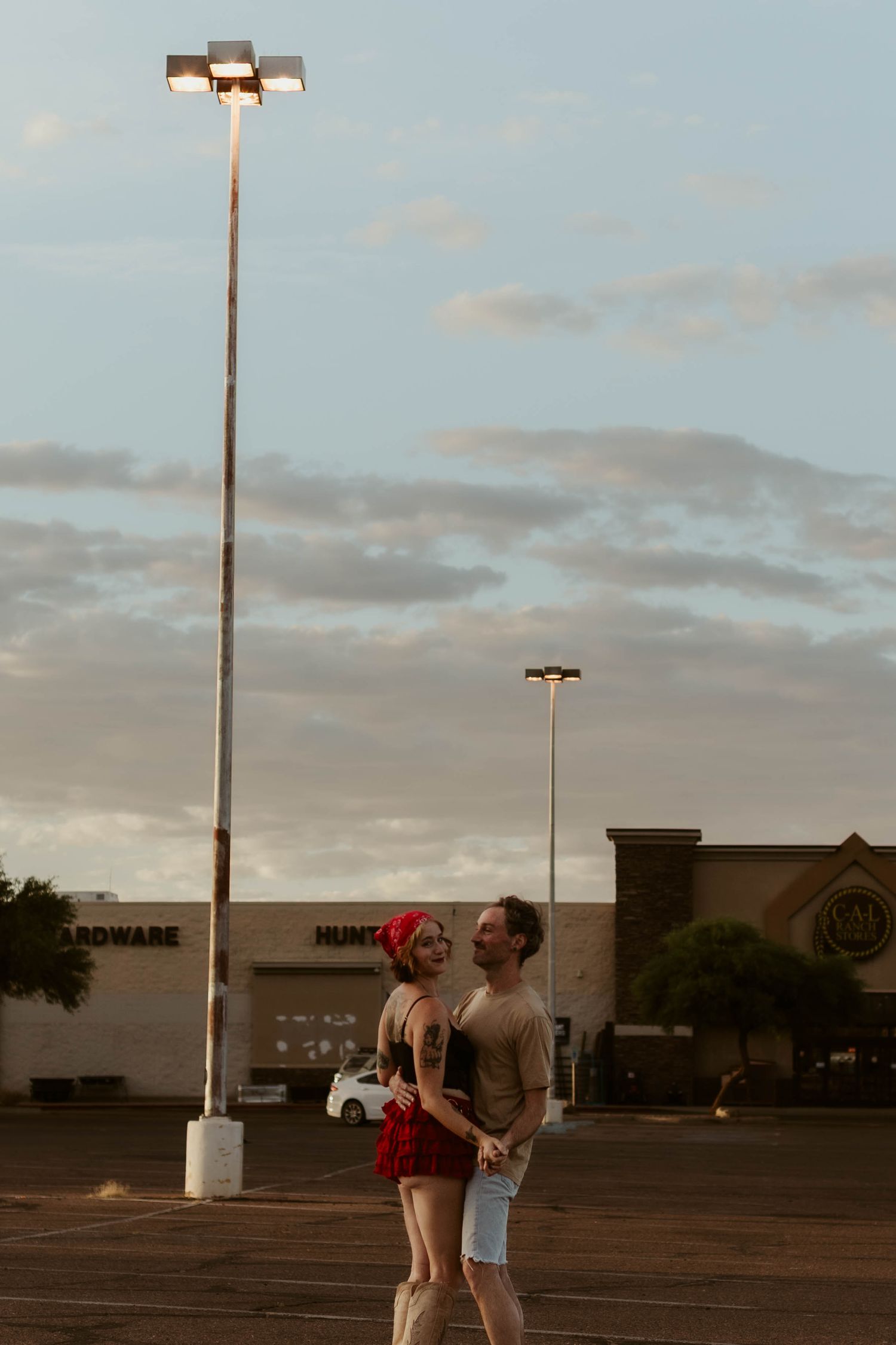 Couple embracing in a parking lot at dusk under a bright street lamp with retail stores in the background.