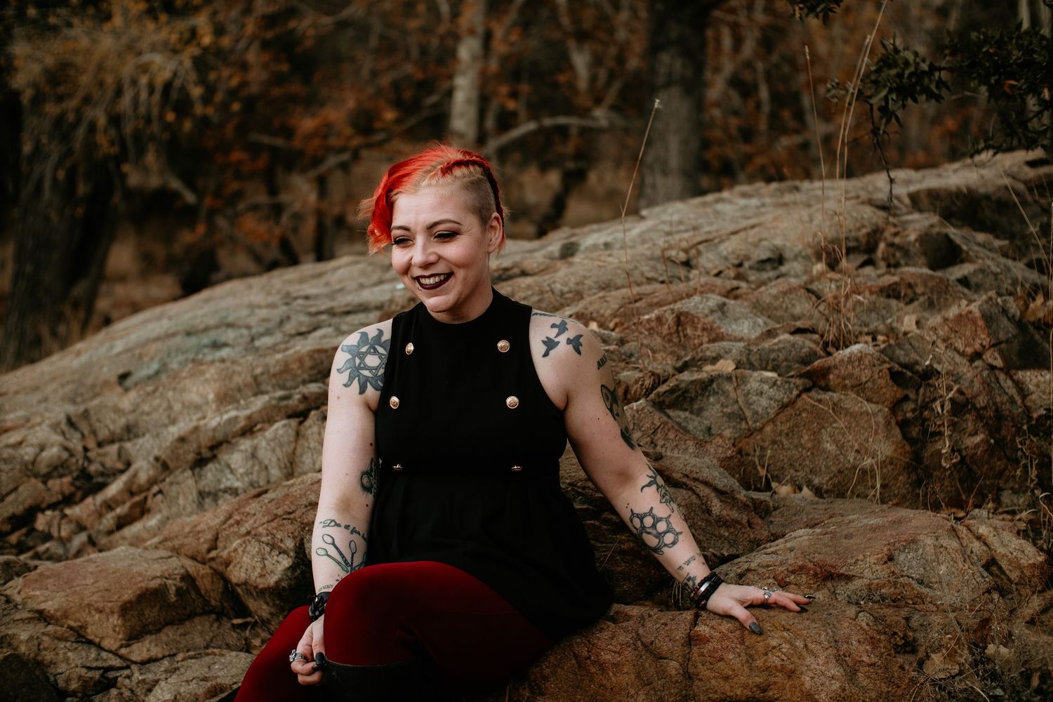 A smiling woman with red hair and tattoos sits on rocks outdoors, wearing a black top and red pants.