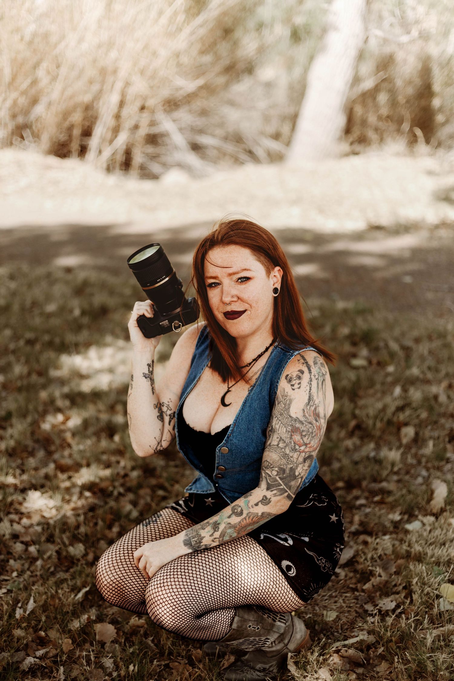 Tattooed redhead woman sitting outdoors holding a DSLR camera, wearing fishnet stockings and denim vest.