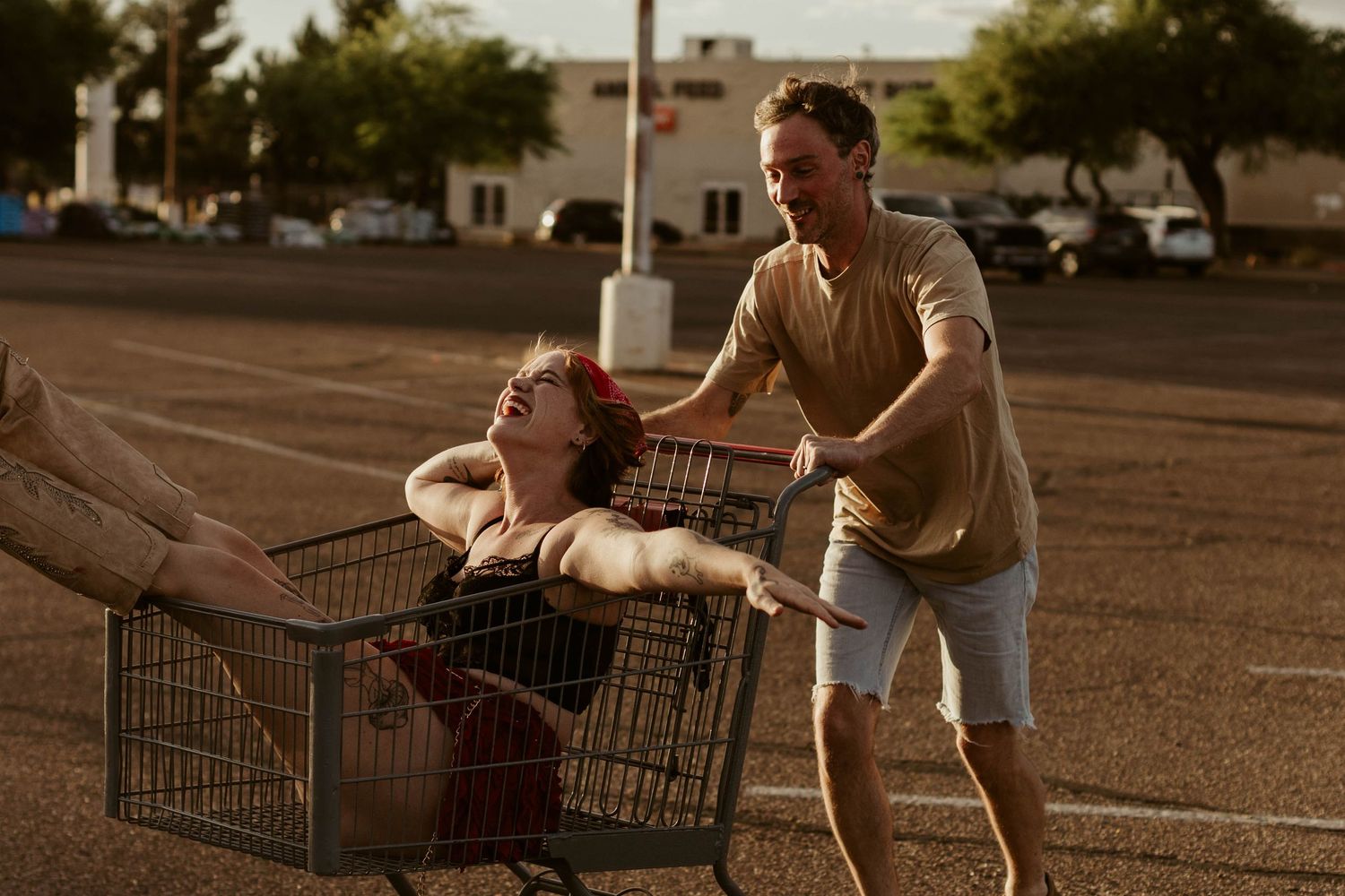 Two friends laugh as one rides in a shopping cart pushed through a sunny parking lot.
