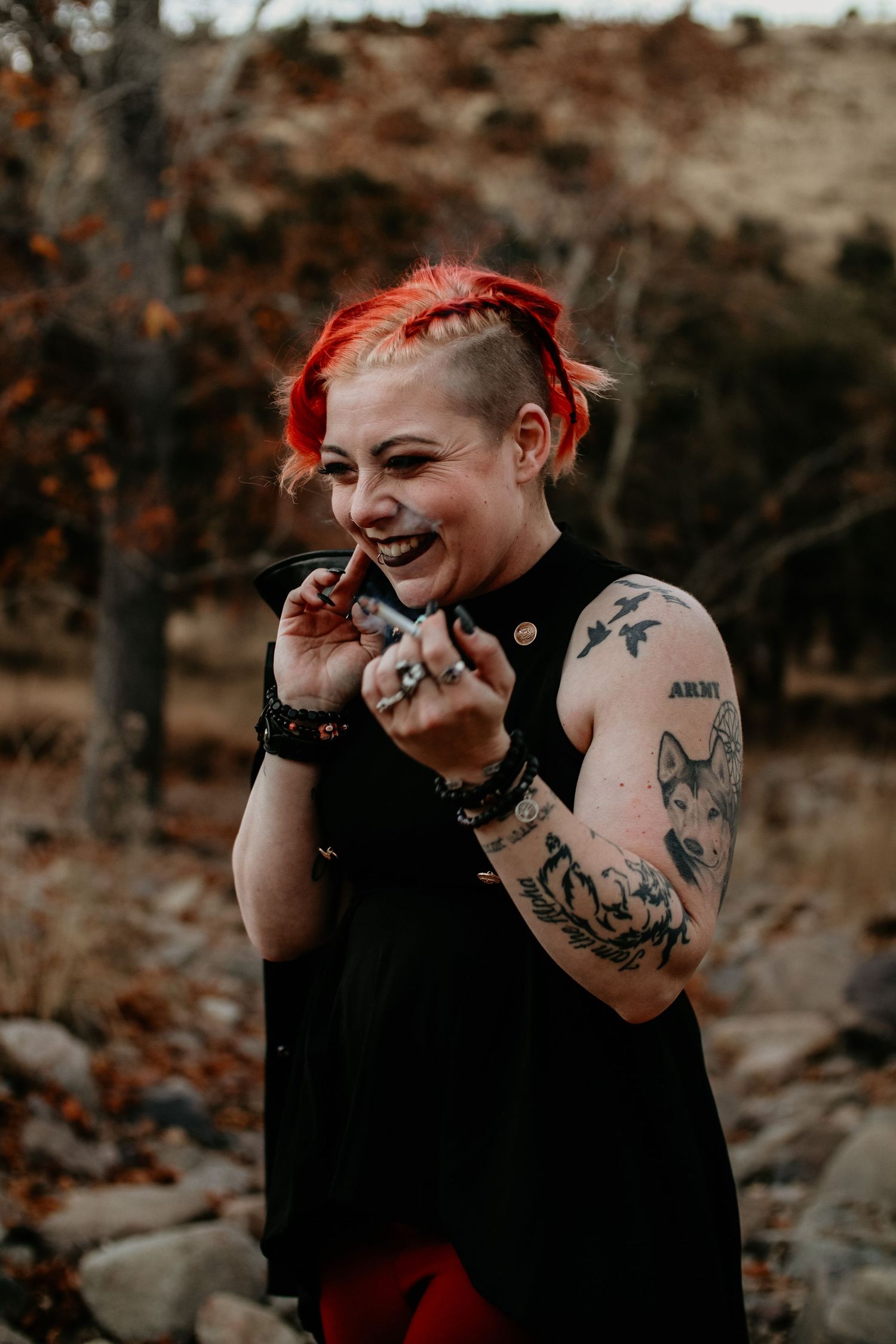 Laughing woman with red hair and tattoos wearing black sleeveless top outdoors in autumn setting.
