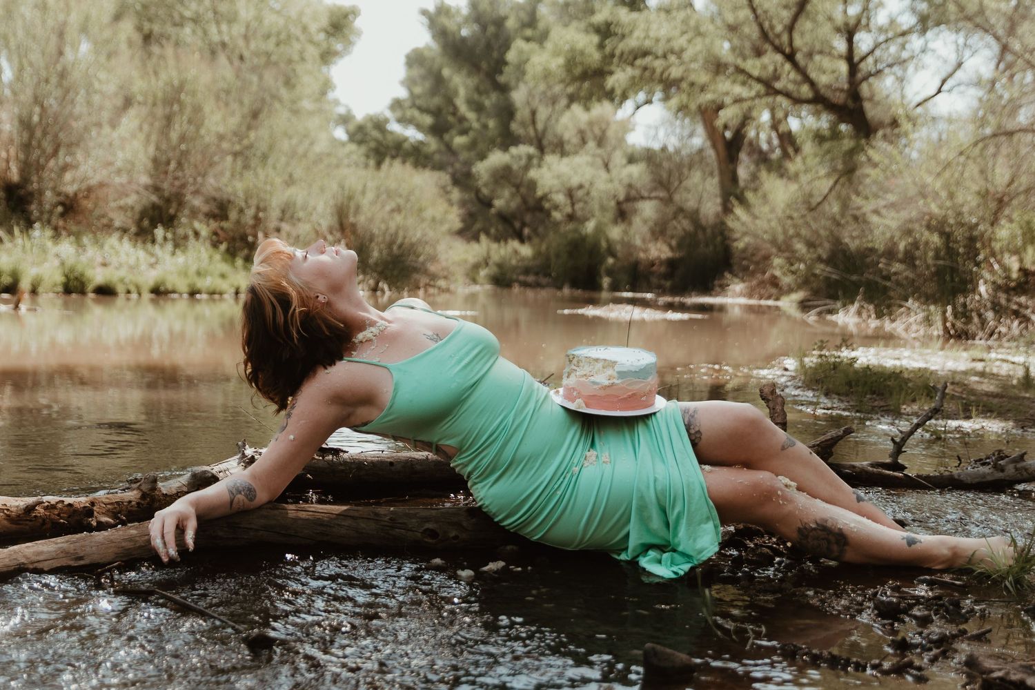 A person in a mint green dress lies gracefully on rocks beside a tranquil stream in a wooded setting.