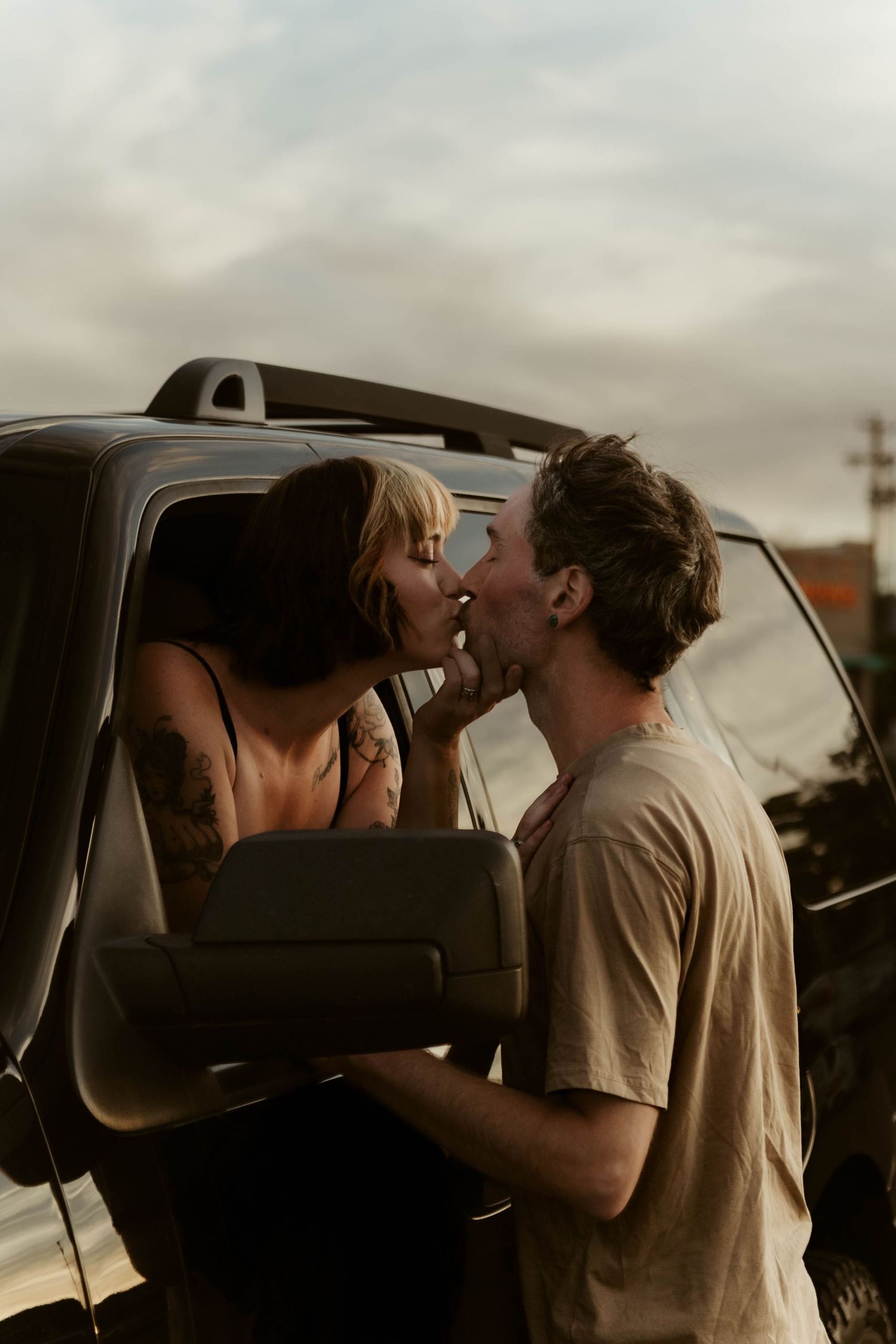 A couple sharing a romantic kiss beside a truck window at dusk, woman with tattoos leaning out toward man.