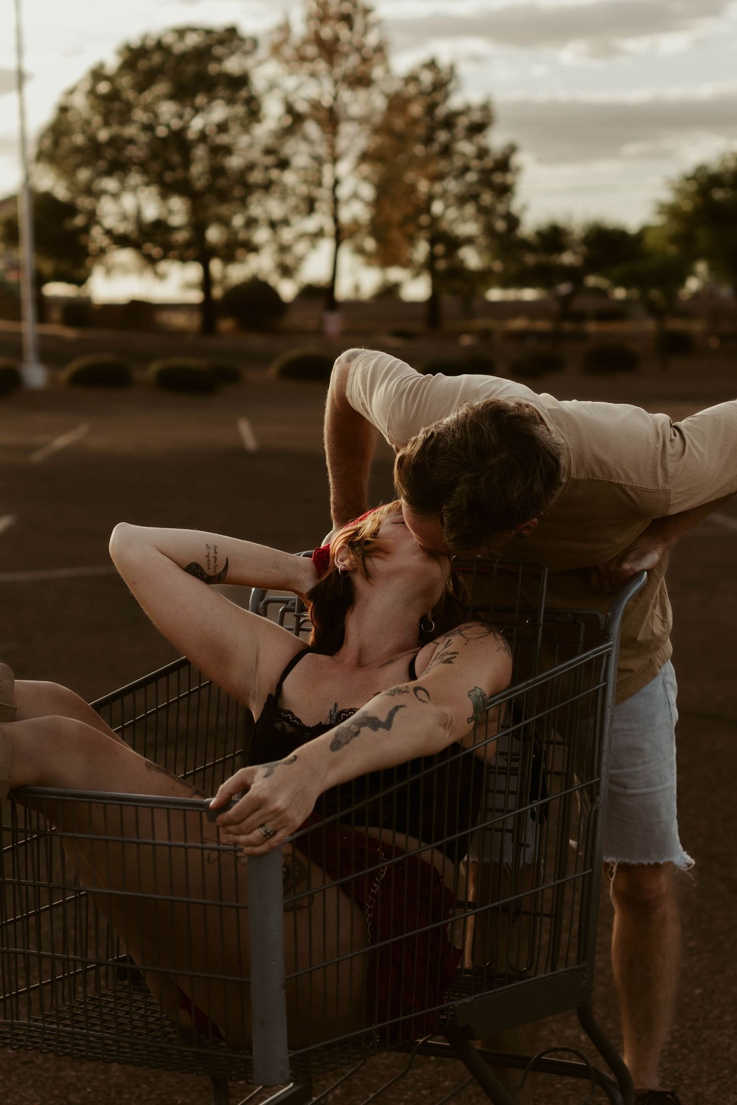 Tattooed woman reclines in shopping cart as man leans in for a kiss in a parking lot at dusk.
