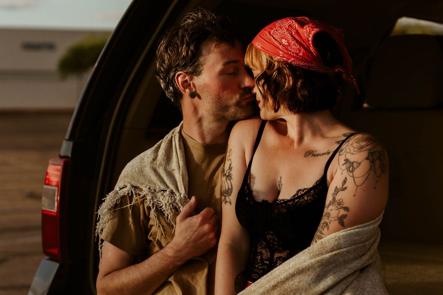 Couple sharing a romantic moment kissing by a car trunk, woman with tattoos and red bandana, golden hour light.