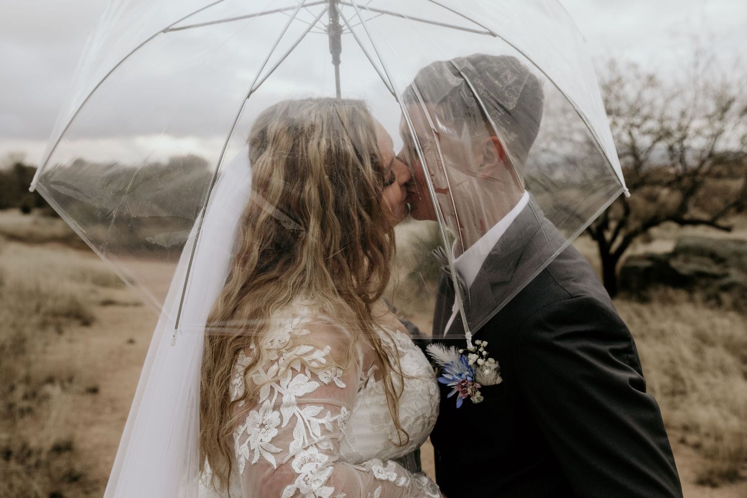 A bride and groom stand close together under a clear umbrella, surrounded by a scenic outdoor landscape.