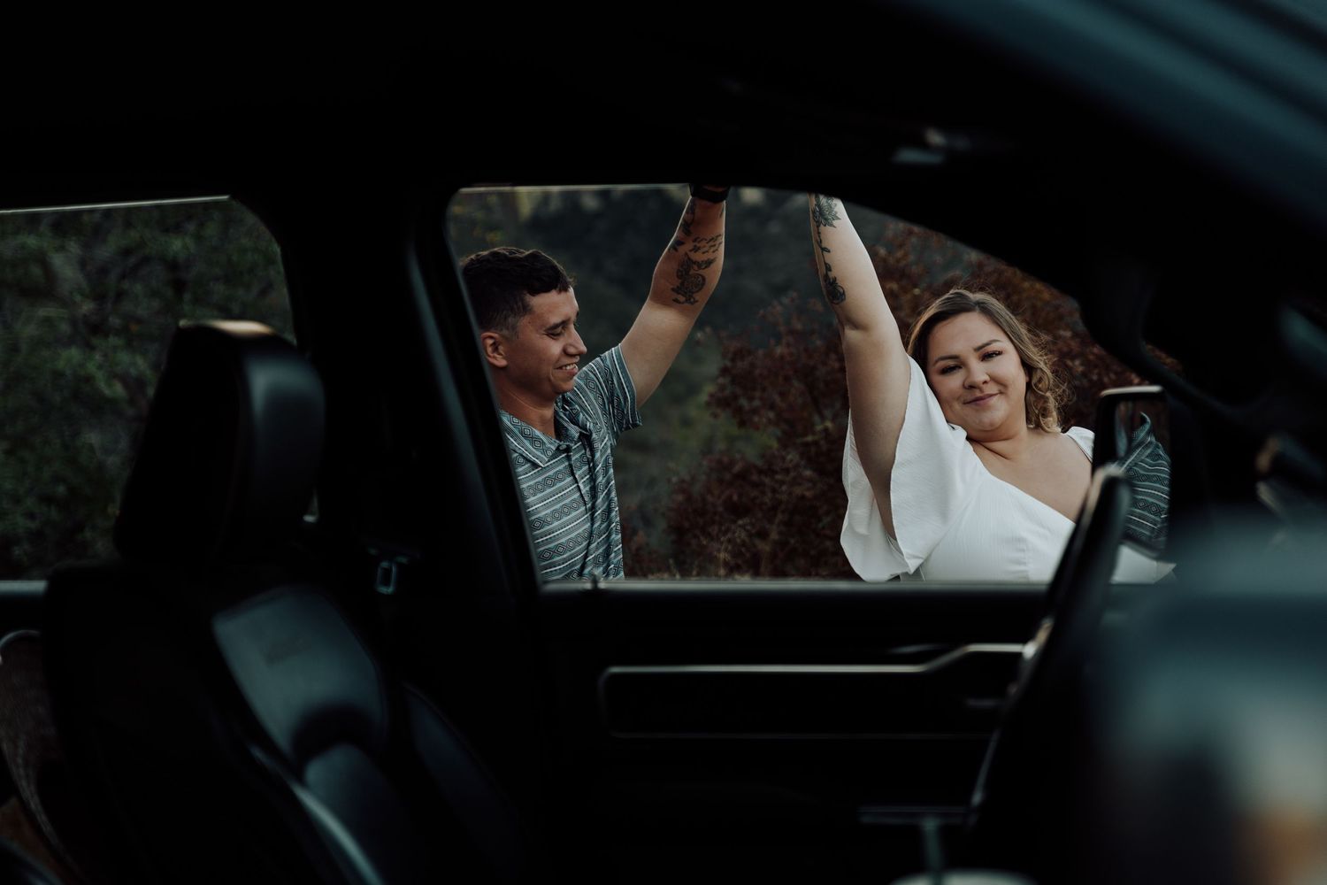A joyful couple dancing and holding hands outside a dark car, surrounded by lush greenery in a romantic outdoor setting.