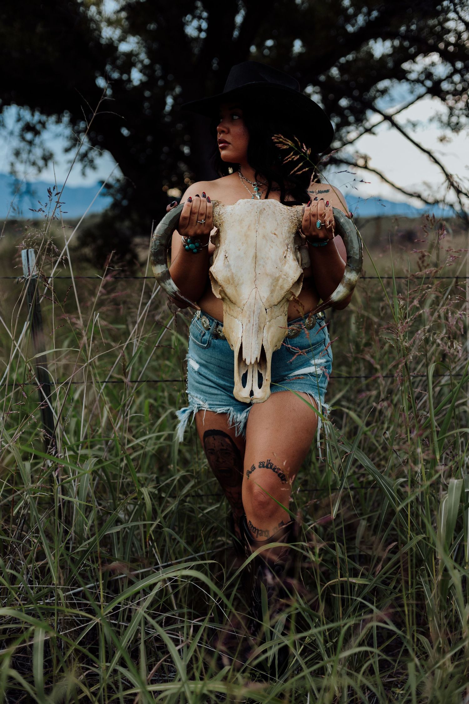 Woman in cowboy hat and denim shorts holding a bull skull in a wild grass field.