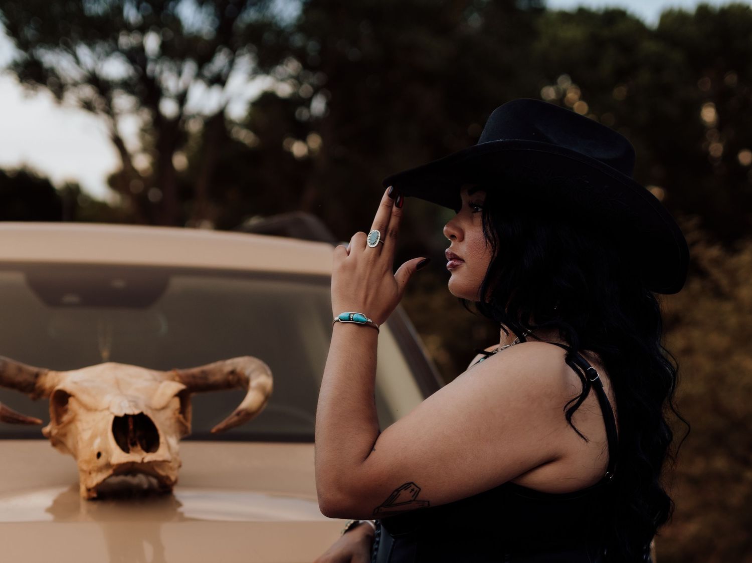 Woman in black cowboy hat poses near vintage car with bull skull in rustic outdoor setting.