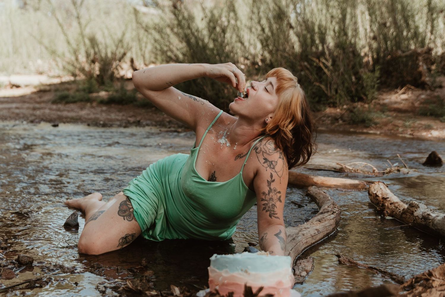 Person in green dress poses artistically near water while covered in body paint and glitter in natural outdoor setting.