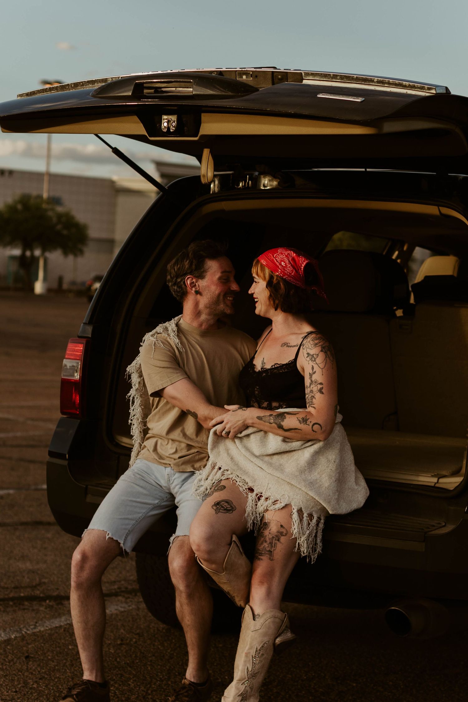 Couple sitting together in open SUV trunk at sunset, woman with red bandana and tattoos sharing romantic moment.