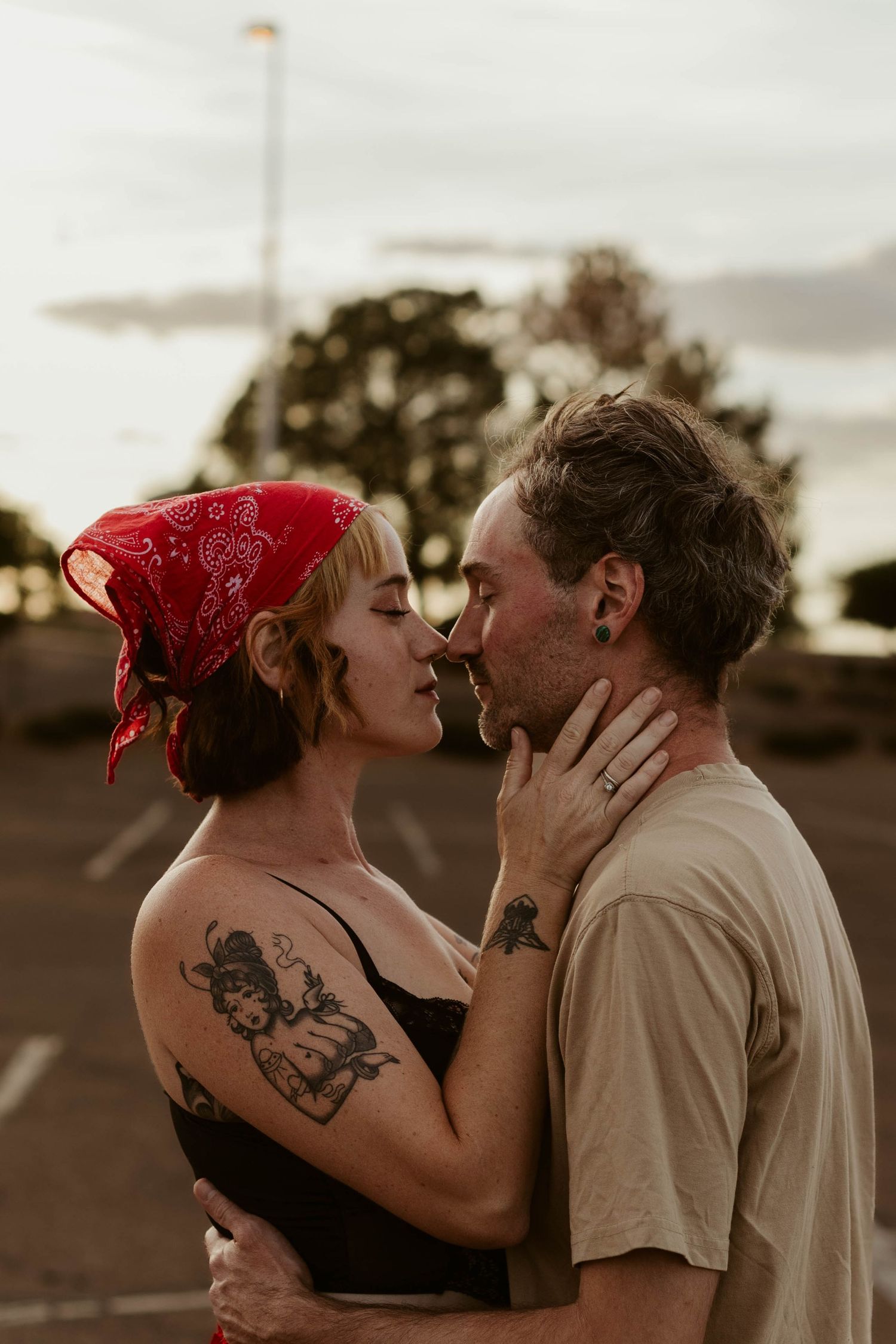 A tattooed couple sharing a romantic moment, nearly kissing outdoors at dusk, woman wearing a red bandana.
