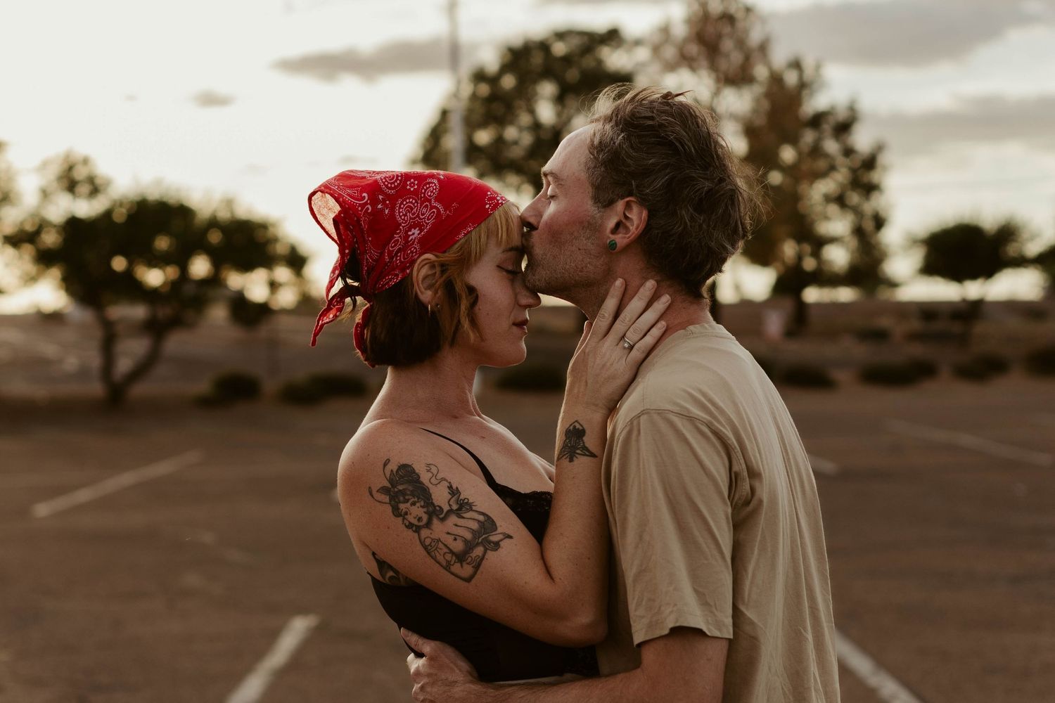 Couple sharing a romantic kiss in a parking lot at sunset, woman wearing red bandana and tattoos.