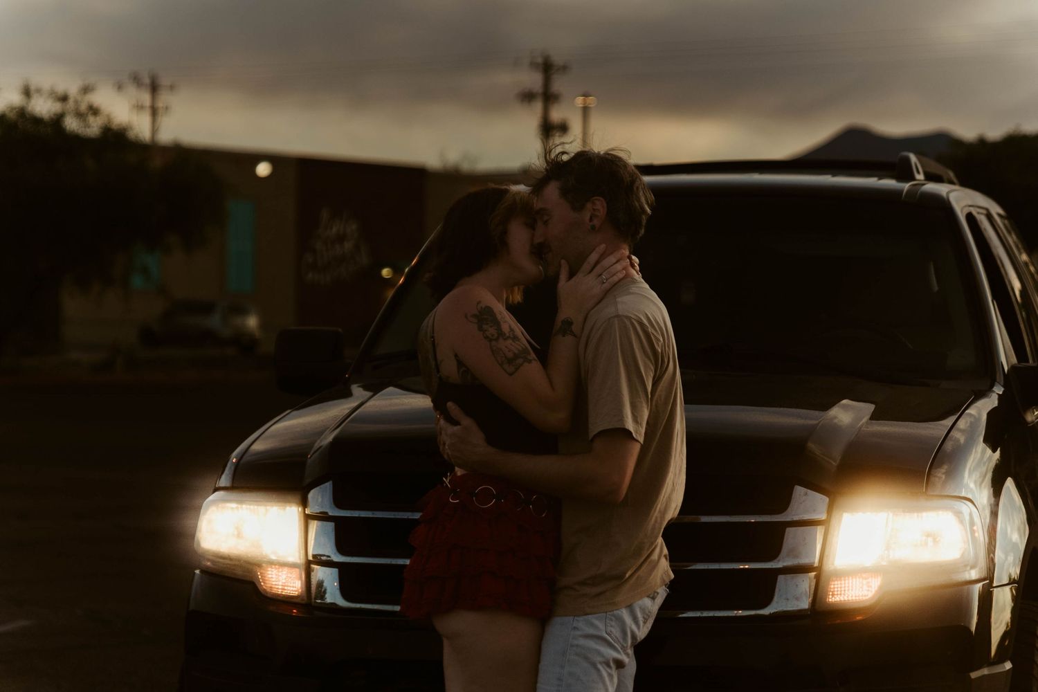 Couple sharing a romantic kiss in front of a black SUV with glowing headlights at dusk.