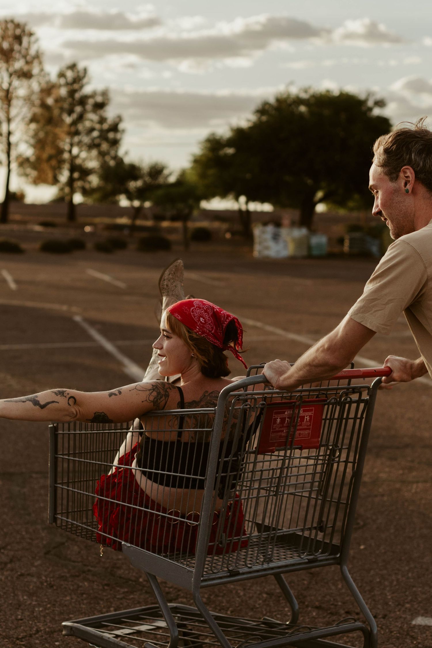 A tattooed woman in a red bandana sits in a shopping cart pushed by a man in a parking lot at sunset.