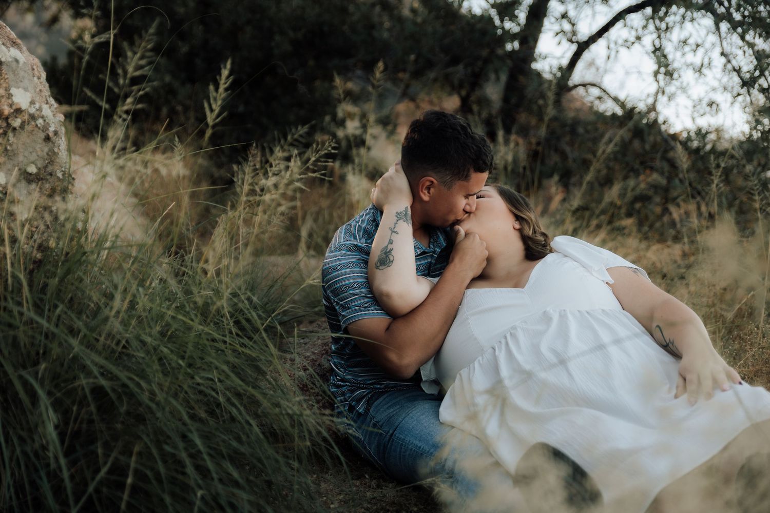 Couple sharing a romantic kiss while sitting in tall grass surrounded by trees in an outdoor engagement photo session.