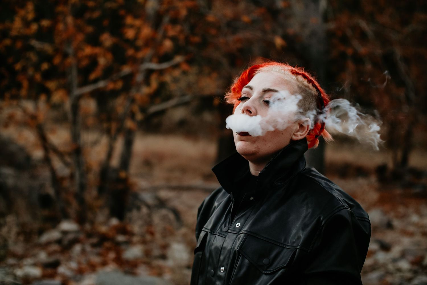 Woman with red hair exhaling smoke outdoors in autumn forest wearing black leather jacket.