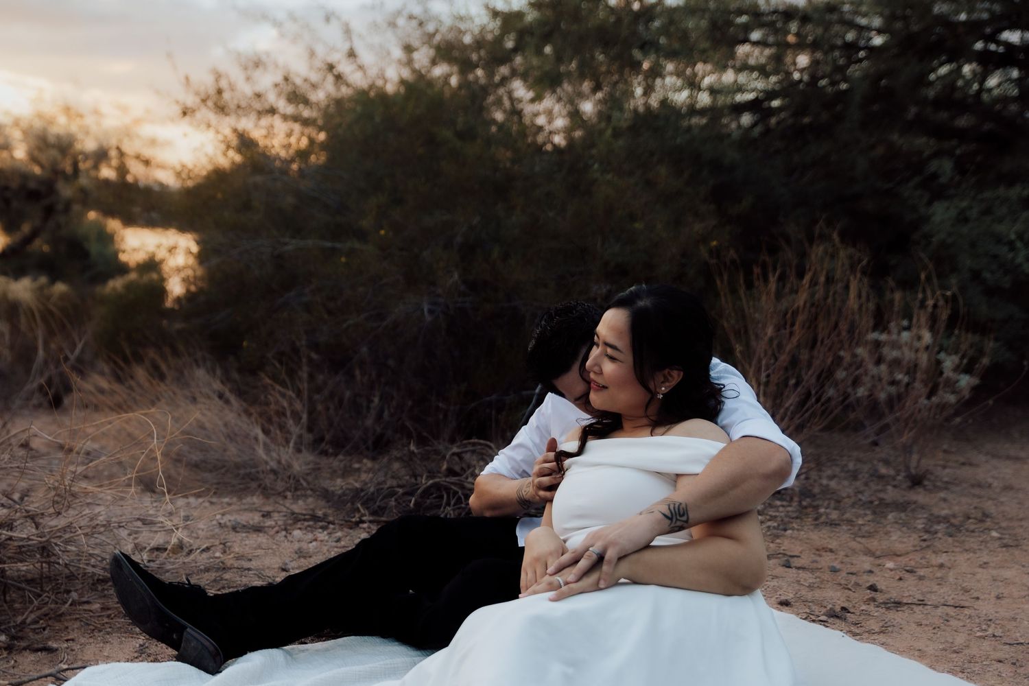 Bride and groom sharing a romantic embrace on desert ground at sunset, surrounded by desert shrubs.