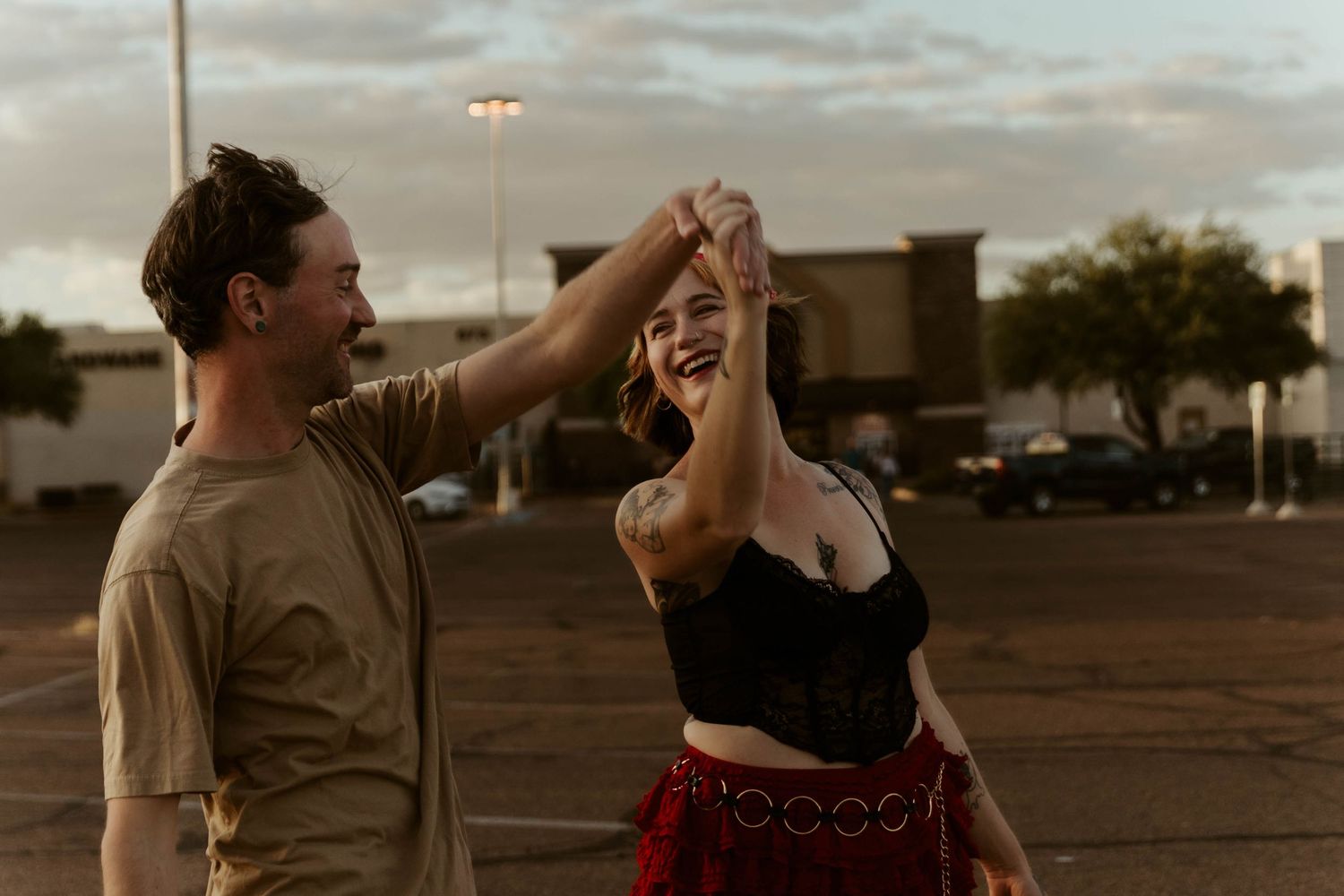 A couple dancing joyfully in a parking lot at dusk, woman in black crop top and red skirt.